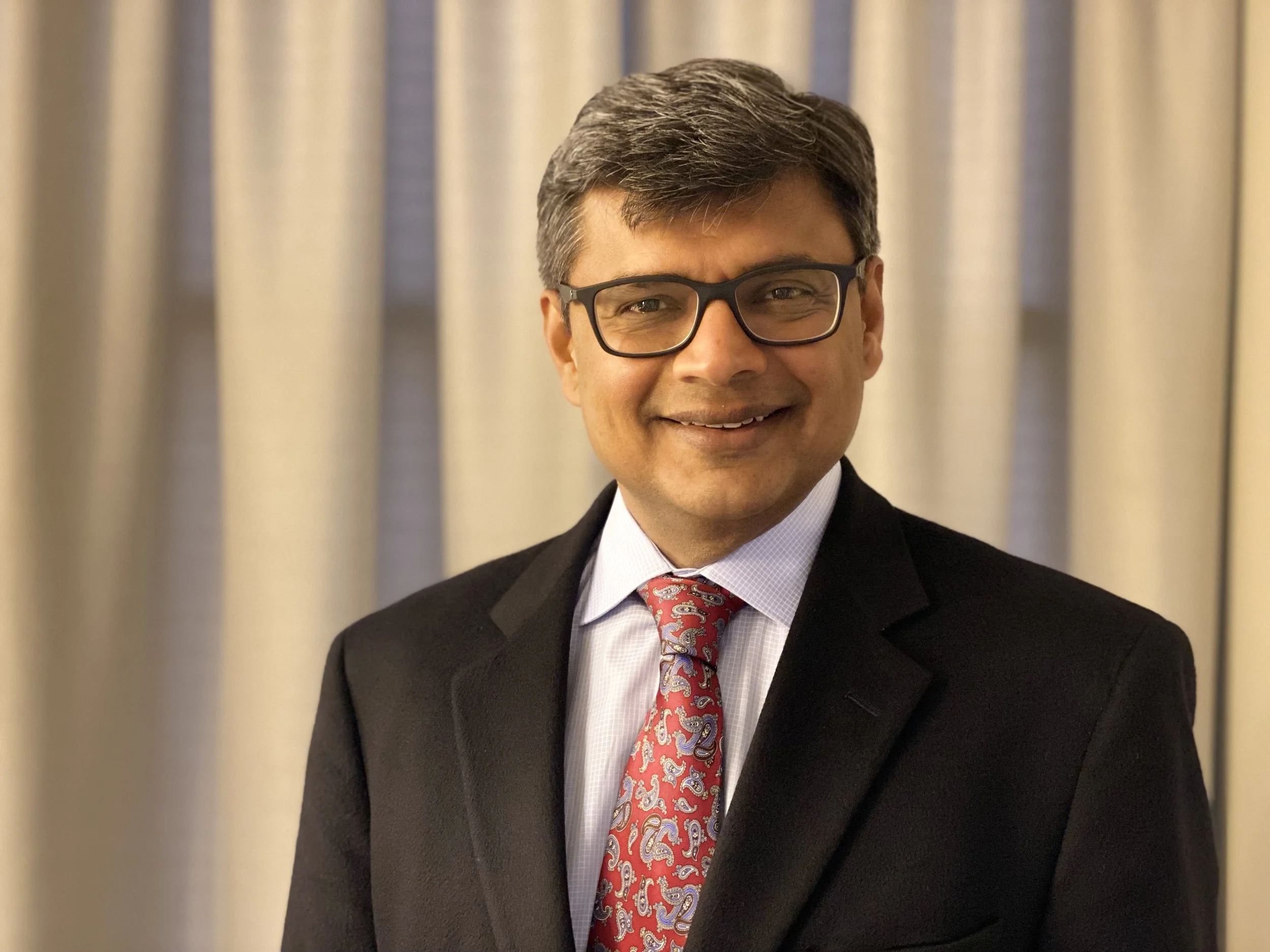 A smiling man in a black suit, white shirt, and red paisley tie, wearing glasses, standing in front of beige curtains.