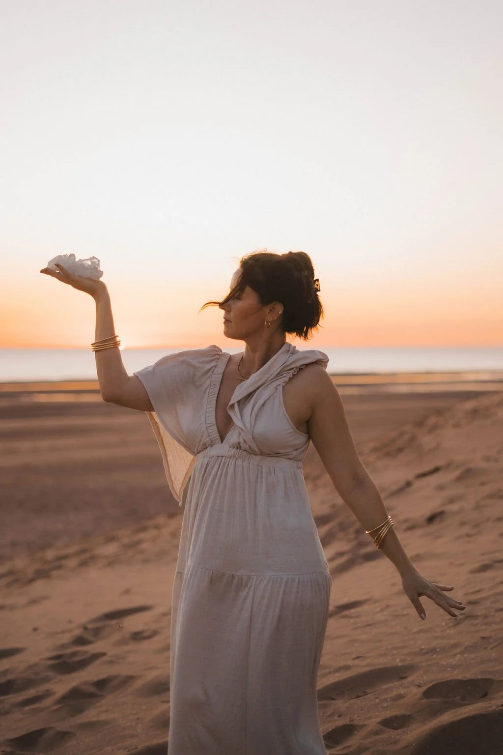 Woman in white dress holding a crumpled paper or cloth on her left hand on a sandy beach during sunset.