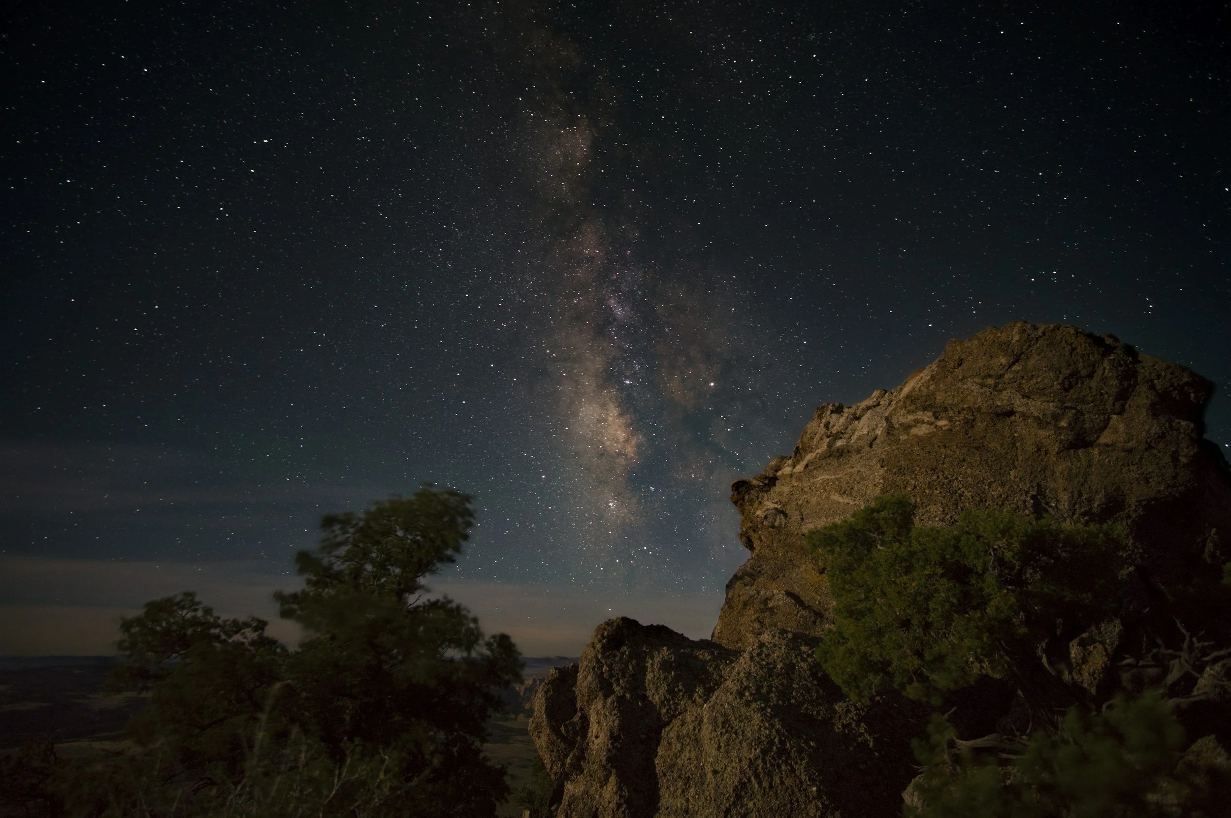 Night sky filled with stars and the Milky Way galaxy, with large rocks and trees in the foreground.
