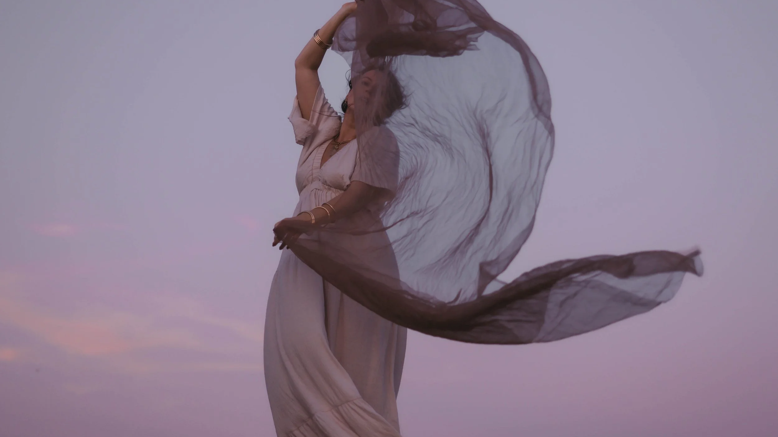 A woman dressed in light pink flowing dress, twirling with a dark, sheer fabric in her hand, against a pastel sky at dusk.