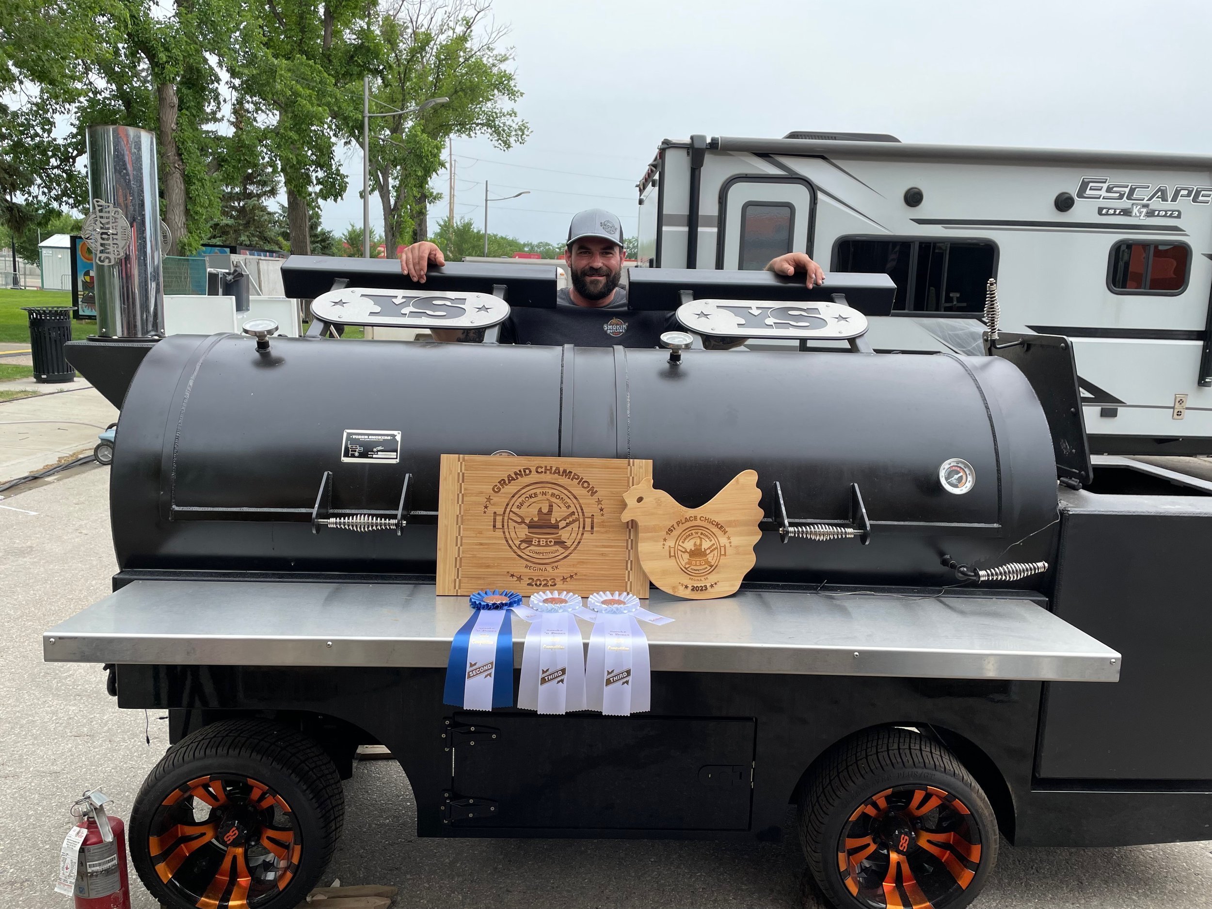 Man behind large outdoor smoker grill with award plaques and ribbons displayed on its base, next to a fire extinguisher, in front of an RV.