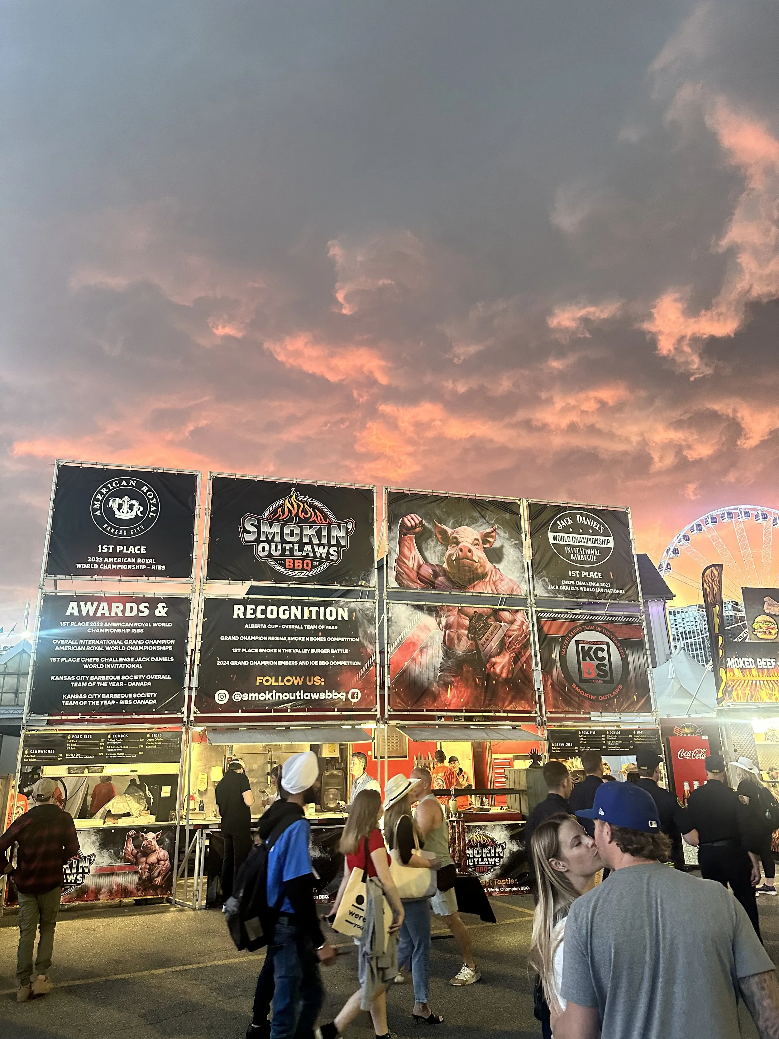 Festival scene with people walking in front of a food stall labeled "Smokin' Outlaws BBQ" displaying awards and recognitions. A colorful, cloudy sunset and a Ferris wheel are visible in the background.