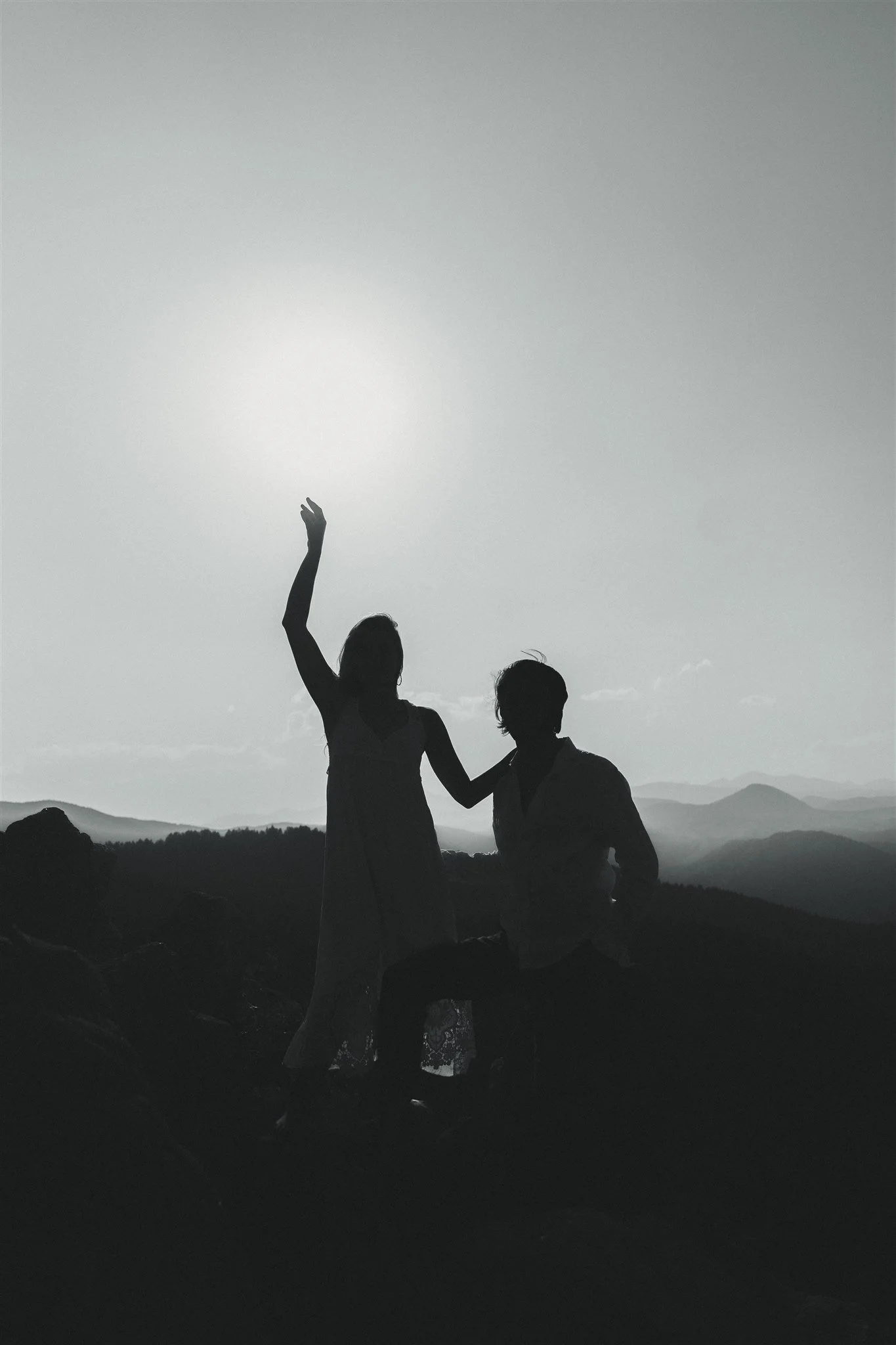 Silhouettes of a couple on a mountain against a bright sky, with mountains in the background