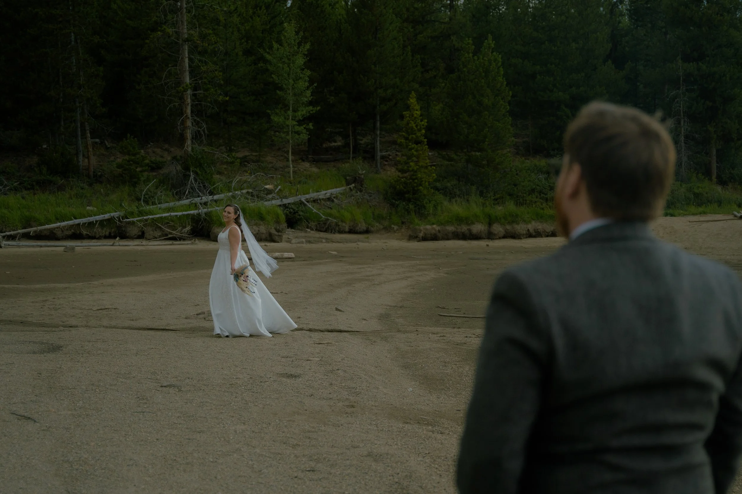 A bride in a white wedding dress holding a bouquet, standing on a sandy beach with a wooded area in the background, while a man in a suit watches her from the foreground.