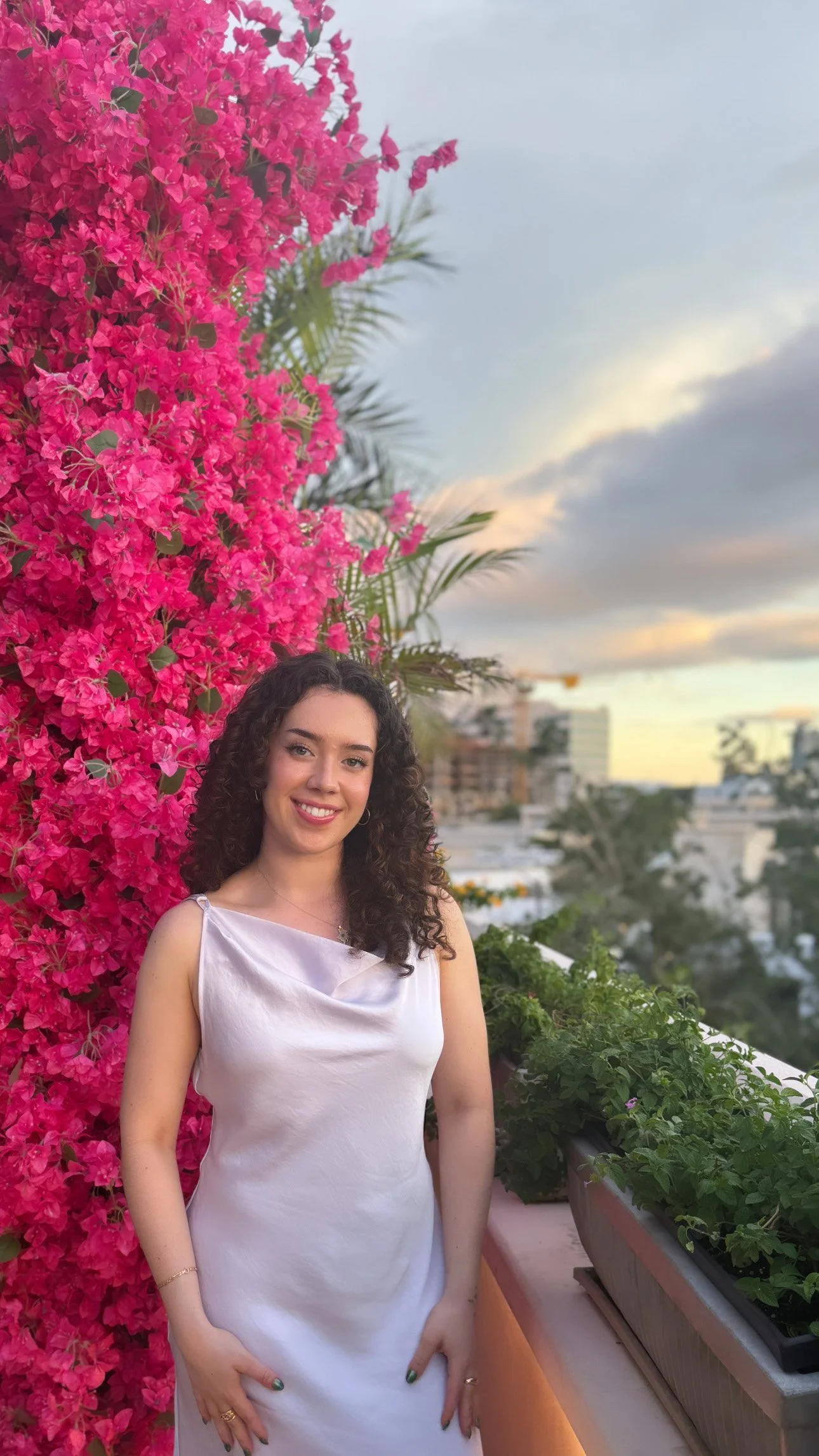 A woman in a satin white dress standing on a balcony at sunset, with pink bougainvillea flowers and a city skyline in the background.
