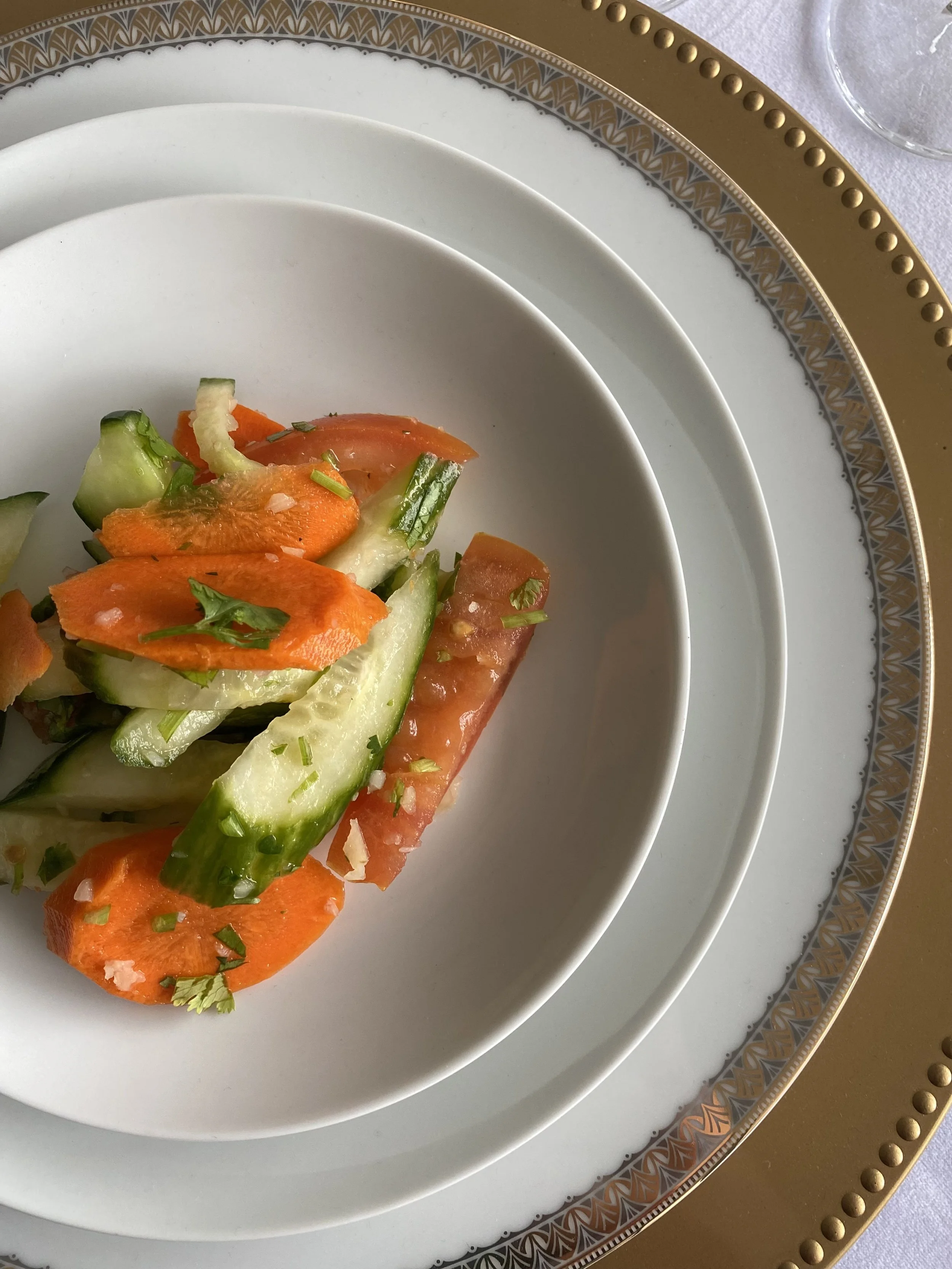 A plate of fresh sliced cucumbers, carrots, and tomatoes garnished with herbs on a white and gold decorative dinnerware