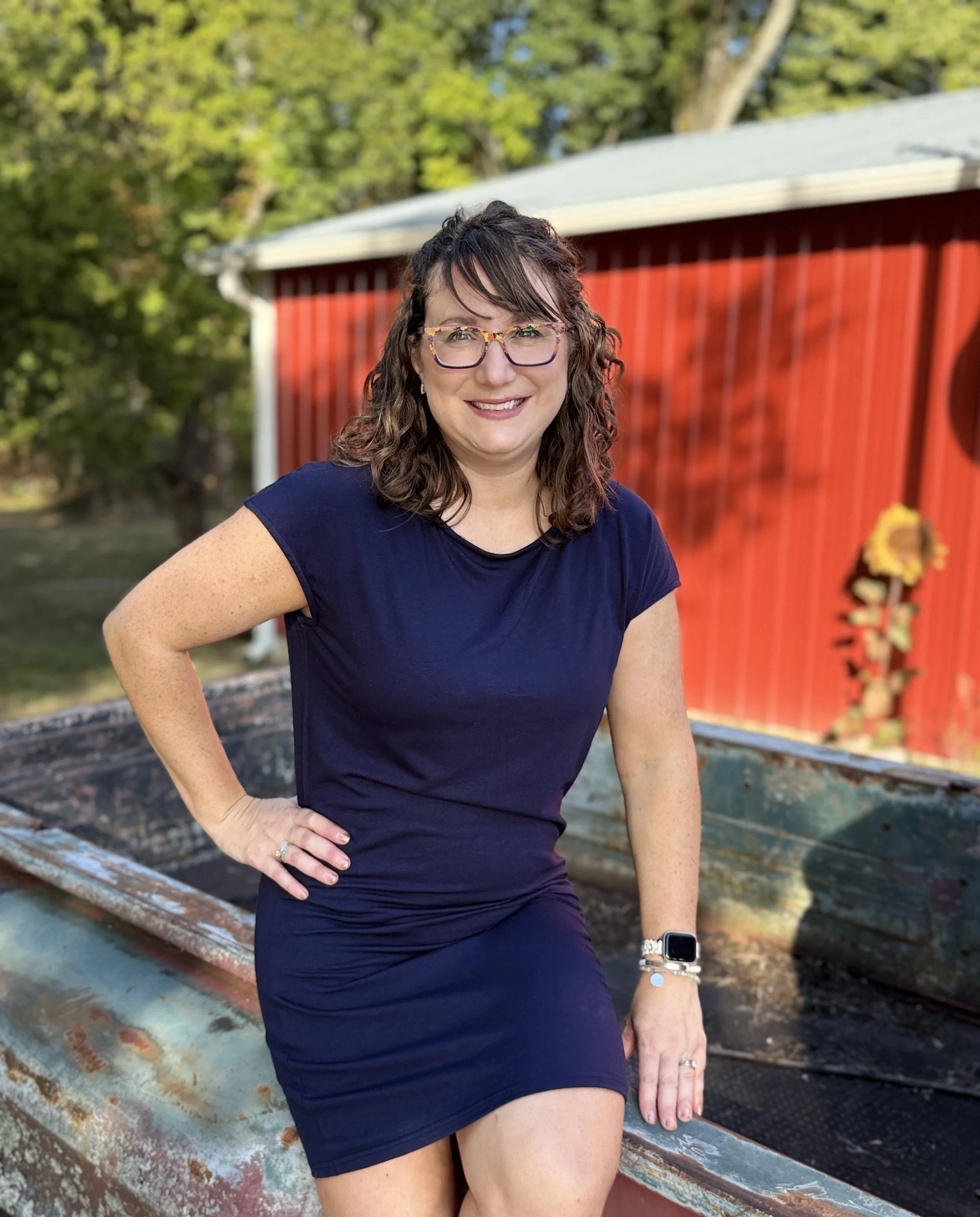 A woman with wavy brown hair and glasses, sitting and smiling on a fall themed antique truck.