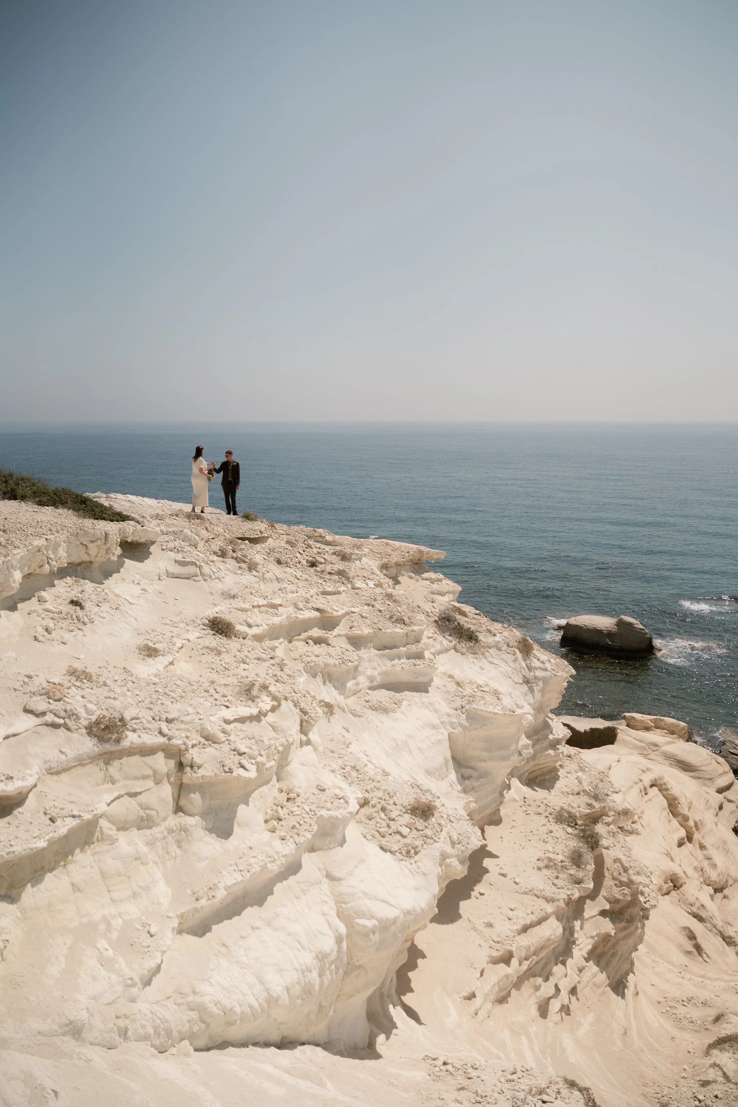 A couple in wedding attire standing on a white rocky cliff by the ocean, exchanging vows or holding hands under clear skies.