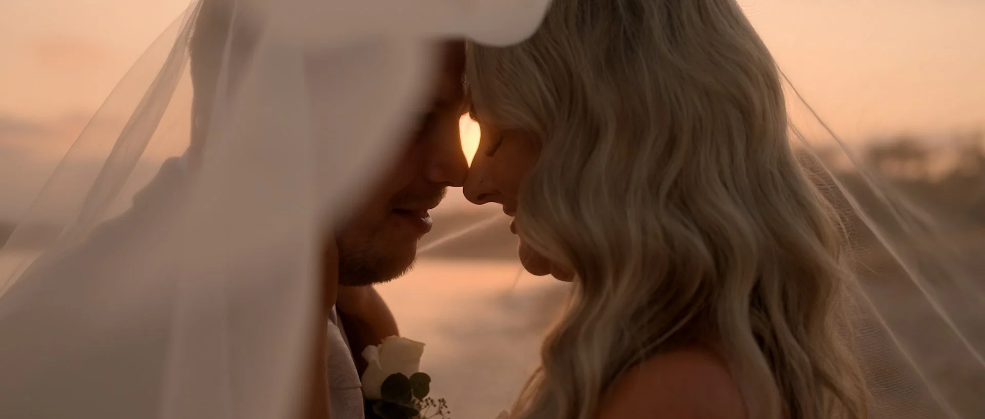 A close-up of a couple touching foreheads under a veil during a wedding, with a cyprus sunset in the background.