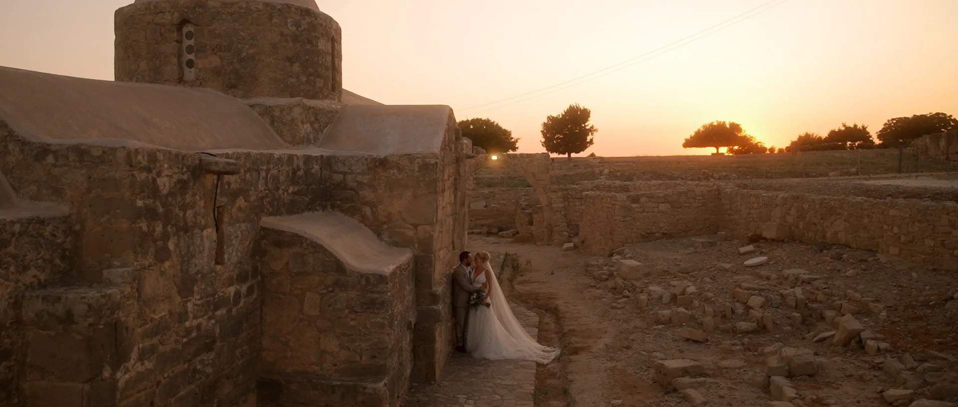 A bride and groom stand close together near liopetro wedding venue at sunset, surrounded by ancient stone ruins and trees in the distance.
