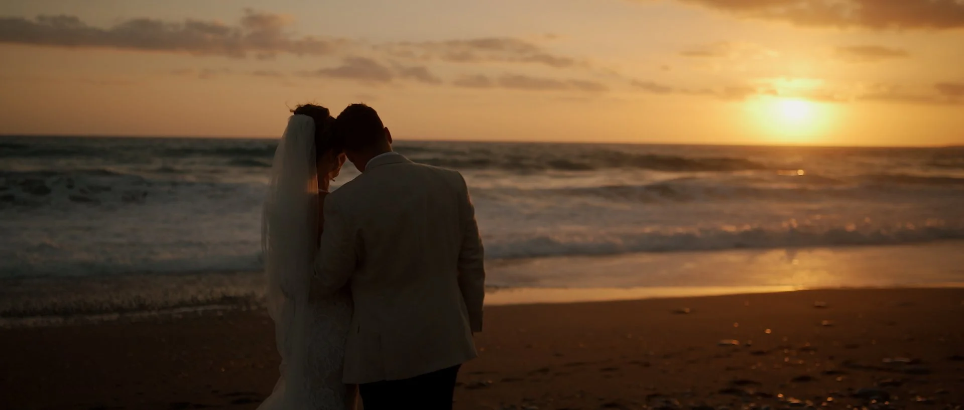 A couple in wedding stands close together on a beach in paphos at sunset, facing the ocean.