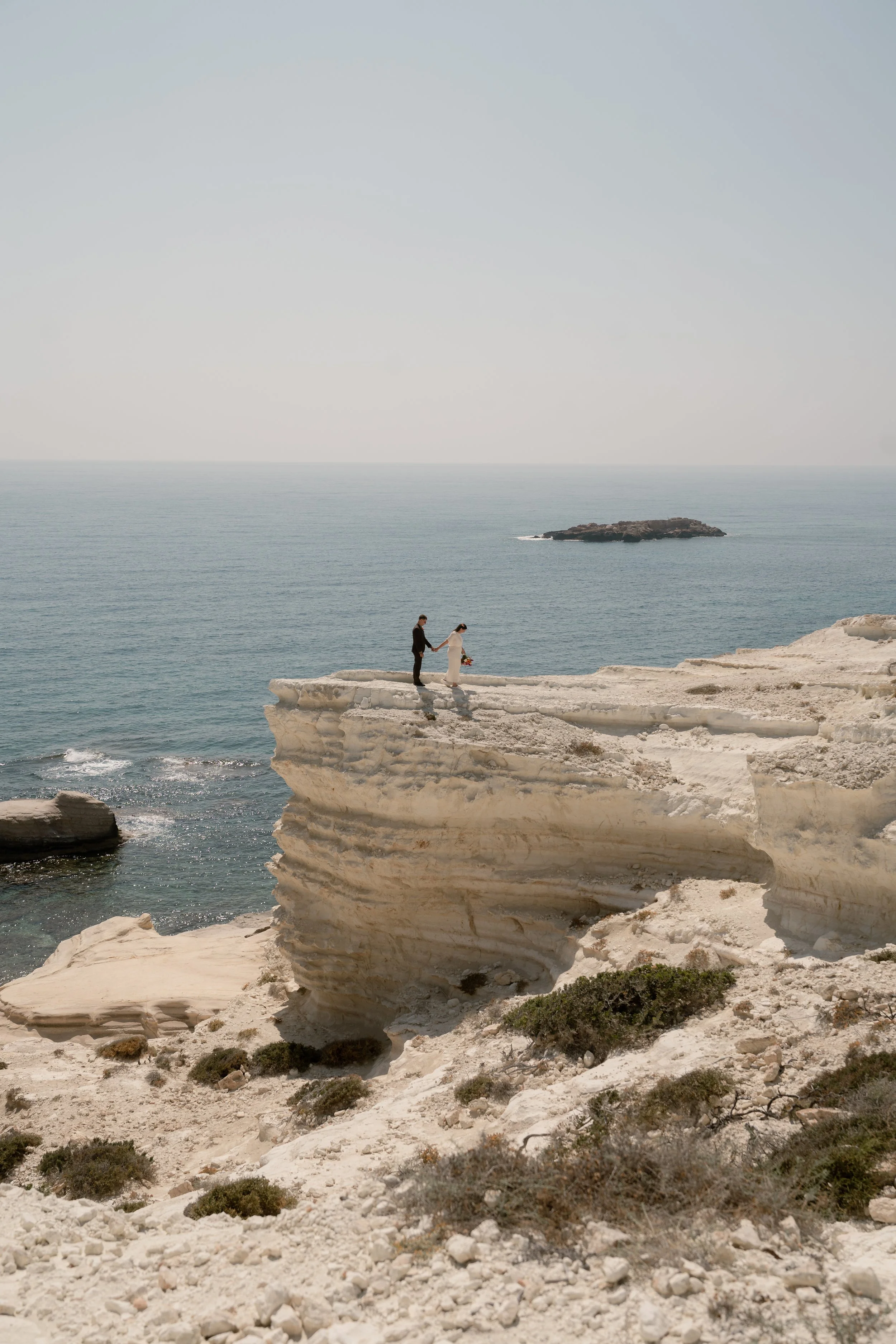 A bride and groom holding hands and walking on a white rocky cliff by the ocean, with the bride holding a bouquet of flowers.