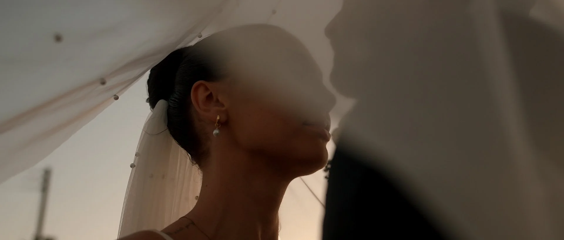 A bride in wedding attire gazing at her groom behind sheer fabric, with her hair in a bun, wearing pearl earrings, in a softly lit room.