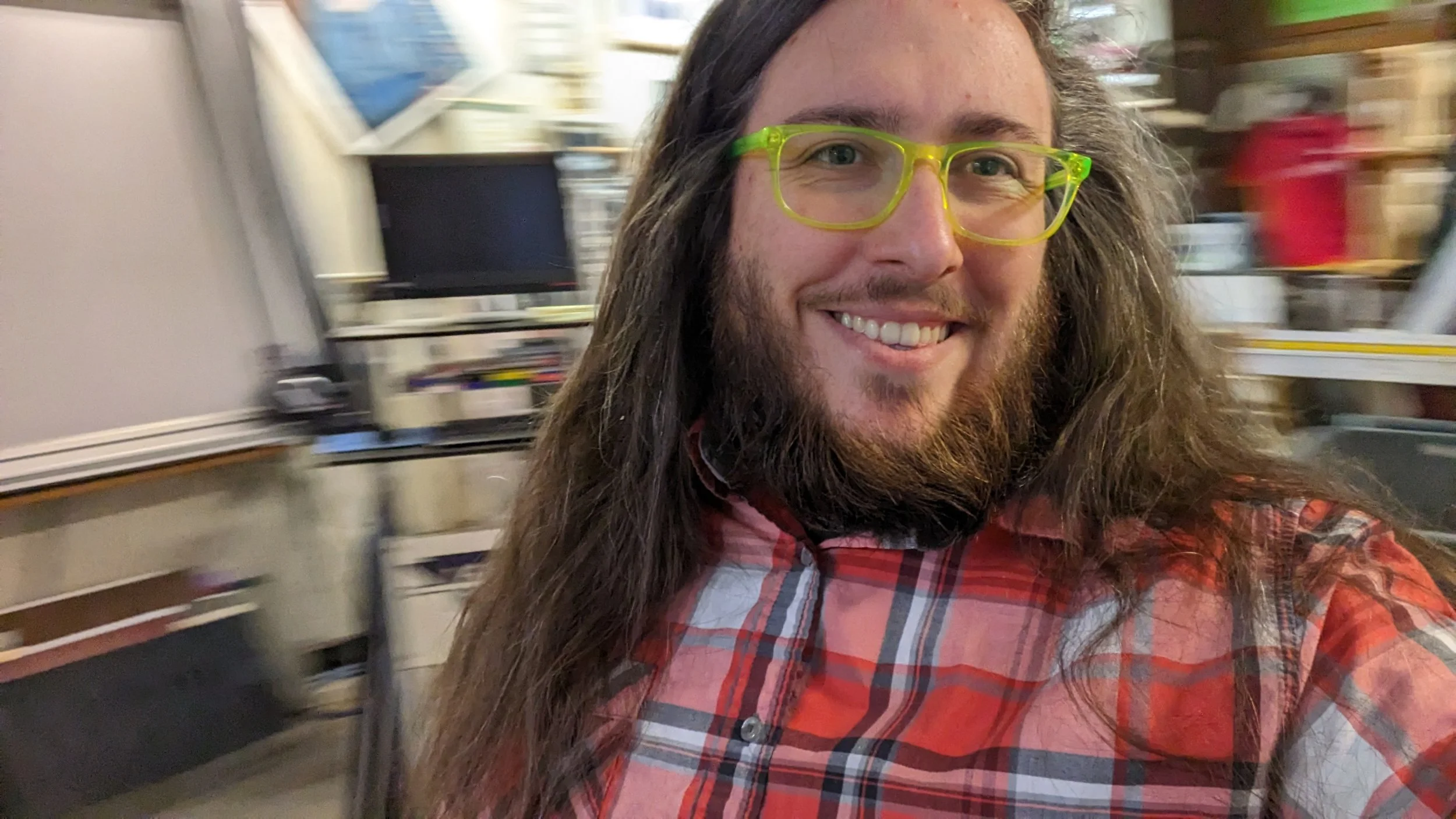 A smiling man with long hair, a beard, and glasses taking a selfie in a cluttered room, wearing a red plaid shirt.