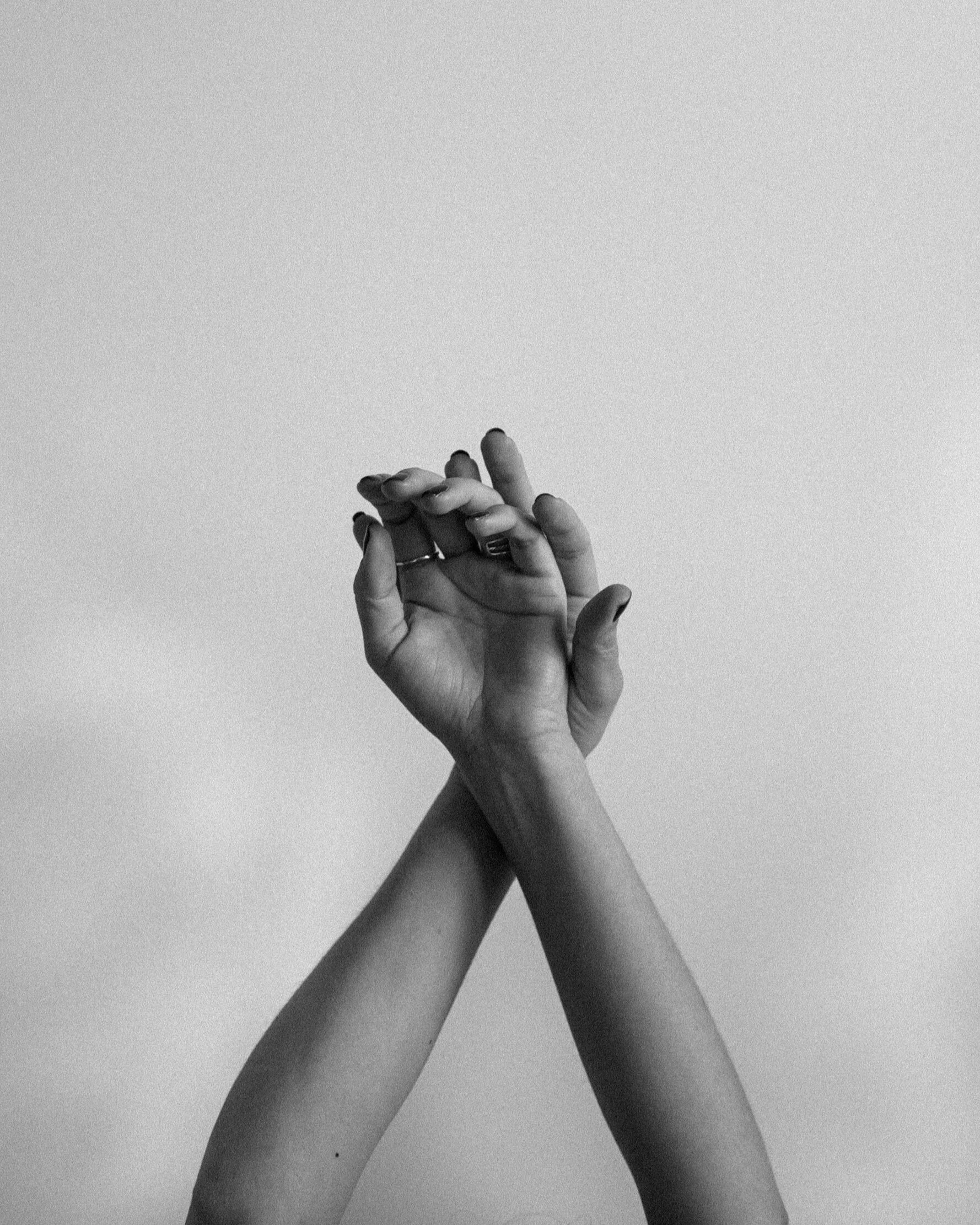 Black and white photo of a person with arms crossed, hands joined, some rings visible on fingers, against a plain background.