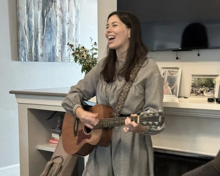 A woman playing an acoustic guitar and singing in a living room with a white fireplace and a TV behind her.