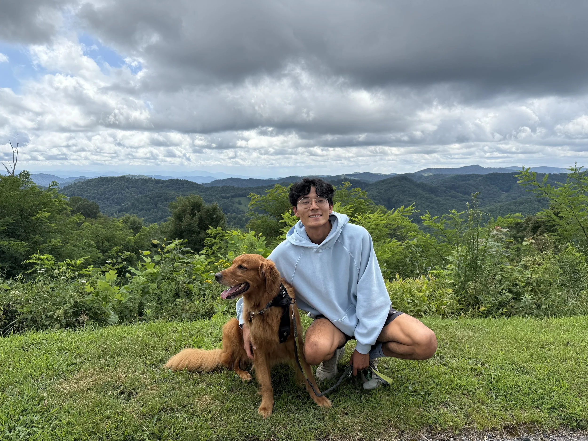 Yudai Koyama in casual clothes and wearing glasses, crouches next to a golden retriever on a trail in a wooded area, both smiling at the camera.
