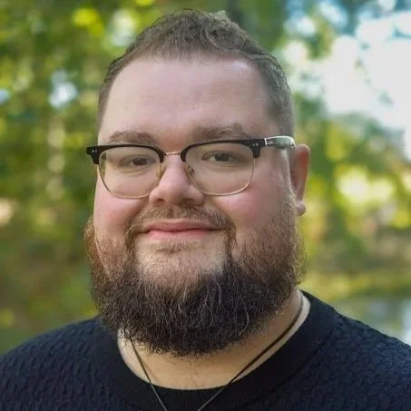 A man with glasses and a beard smiling in front of a modern wall decorated with black and gold circular accents.