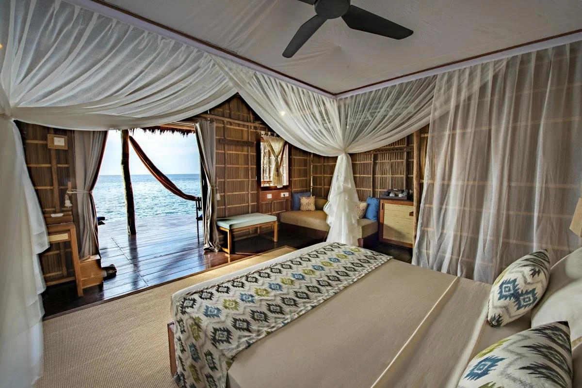 Bedroom with mosquito net canopy, overlooking ocean through open sliding doors, with wooden decor and bedding with geometric patterns.