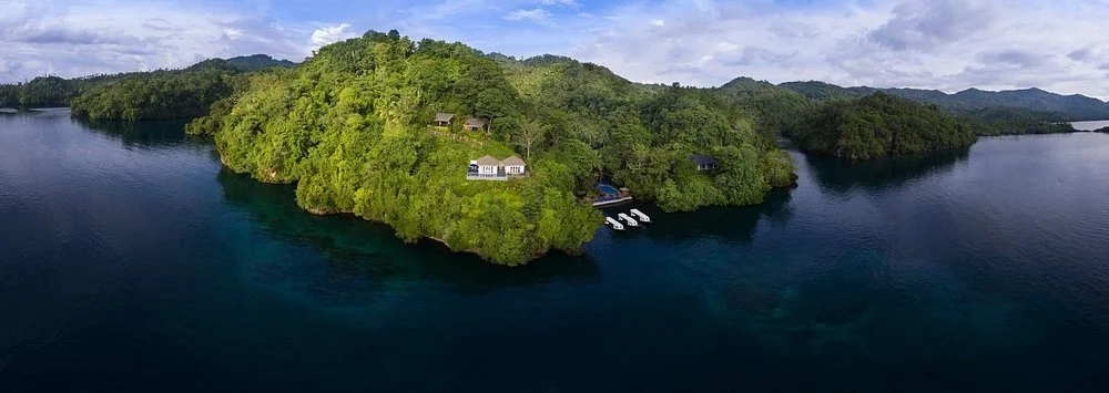 Lembeh Resort - Aerial view of a lush green island surrounded by a calm lake with a few houses and boat docks.