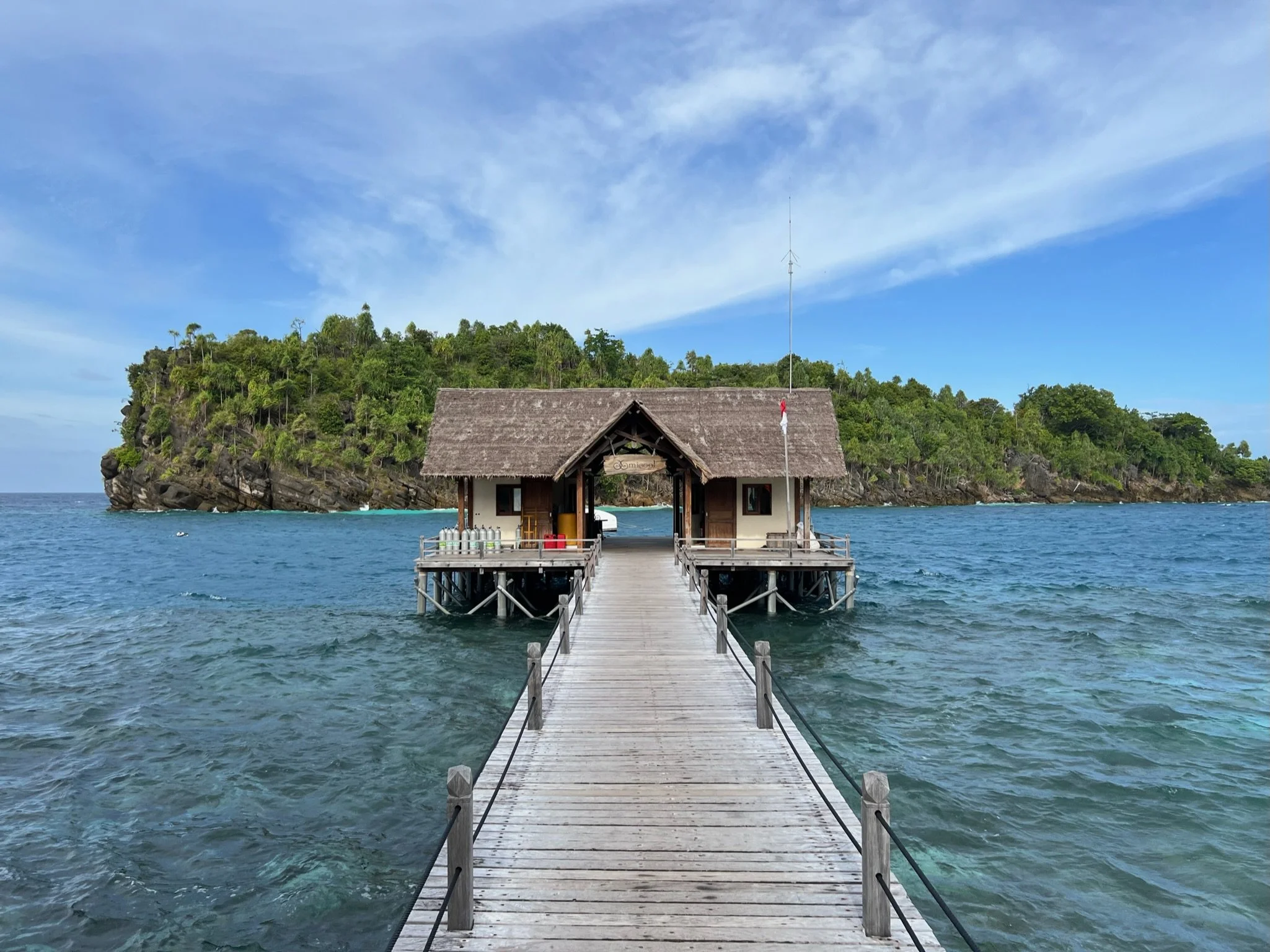 A wooden pier extends over turquoise ocean waters towards a thatched-roof hut on stilts, with a lush green island in the background under a partly cloudy sky.
