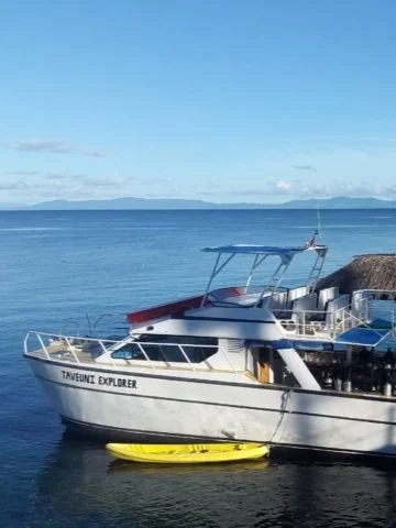 Boat named Taveunui Explorer docked in calm water with a yellow kayak underneath it, with a rocky shoreline and distant land in the background.