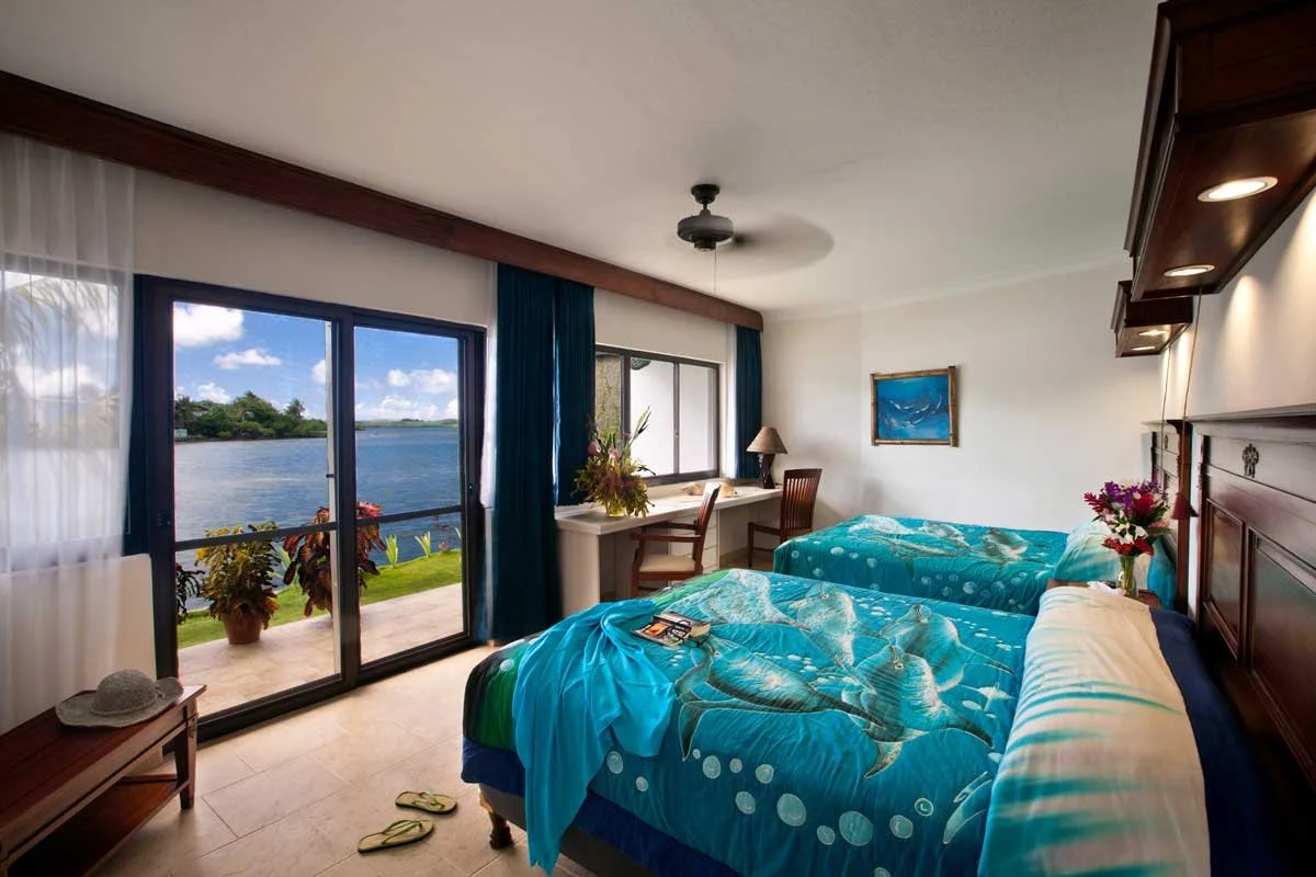 A bedroom with two twin beds featuring dolphin-themed bedding, a sliding glass door with a view of a lake, and tropical plants outside.