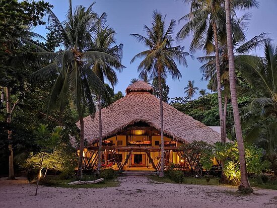 A tropical hut with a thatched roof surrounded by palm trees and lush greenery at sunset