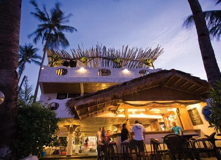 Outdoor bar with thatched roof, illuminated, surrounded by palm trees, with people sitting and socializing during dusk.