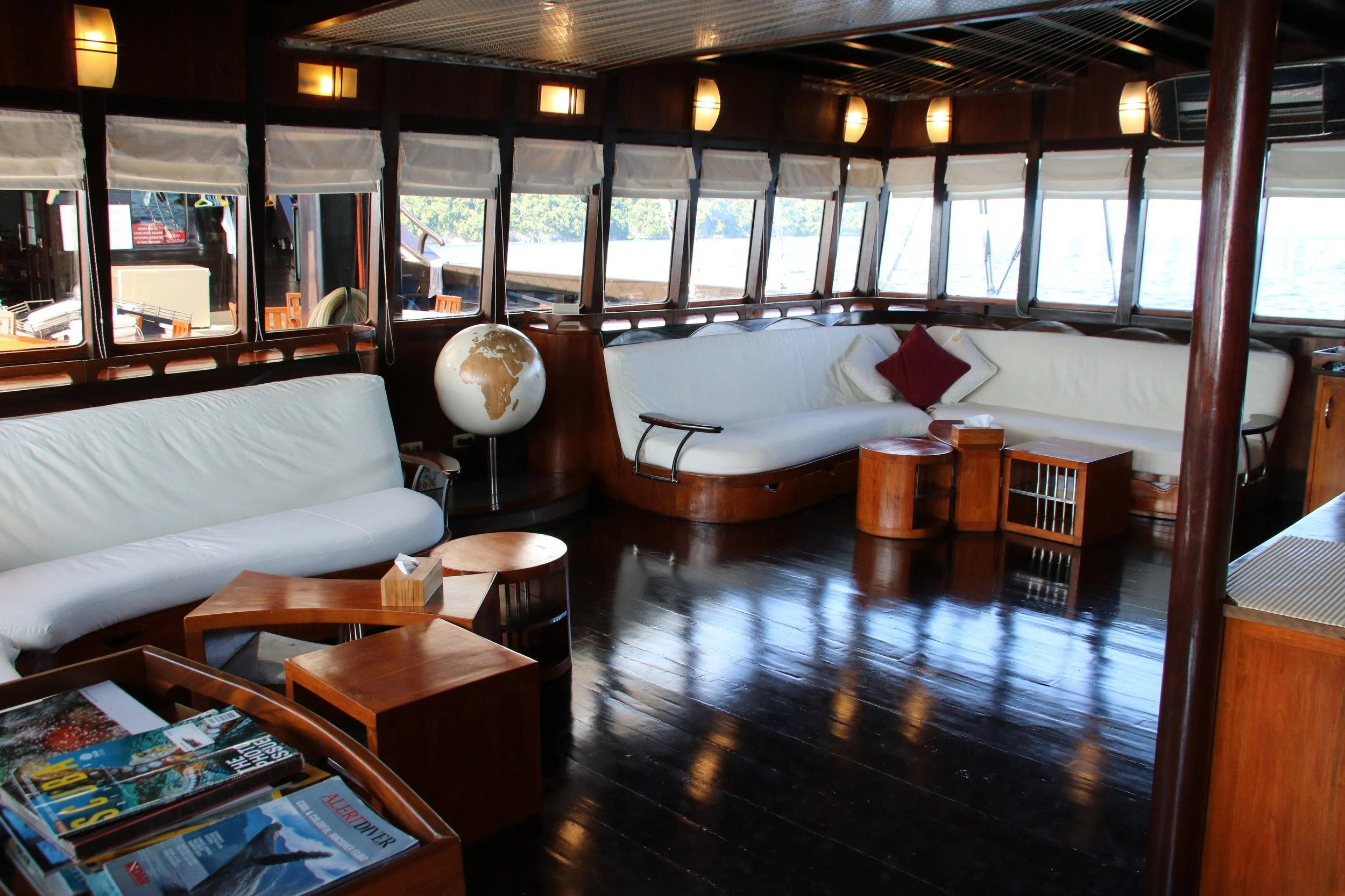 Interior of a boat's lounge with white sofas, wooden tables, a globe, and windows overlooking water.