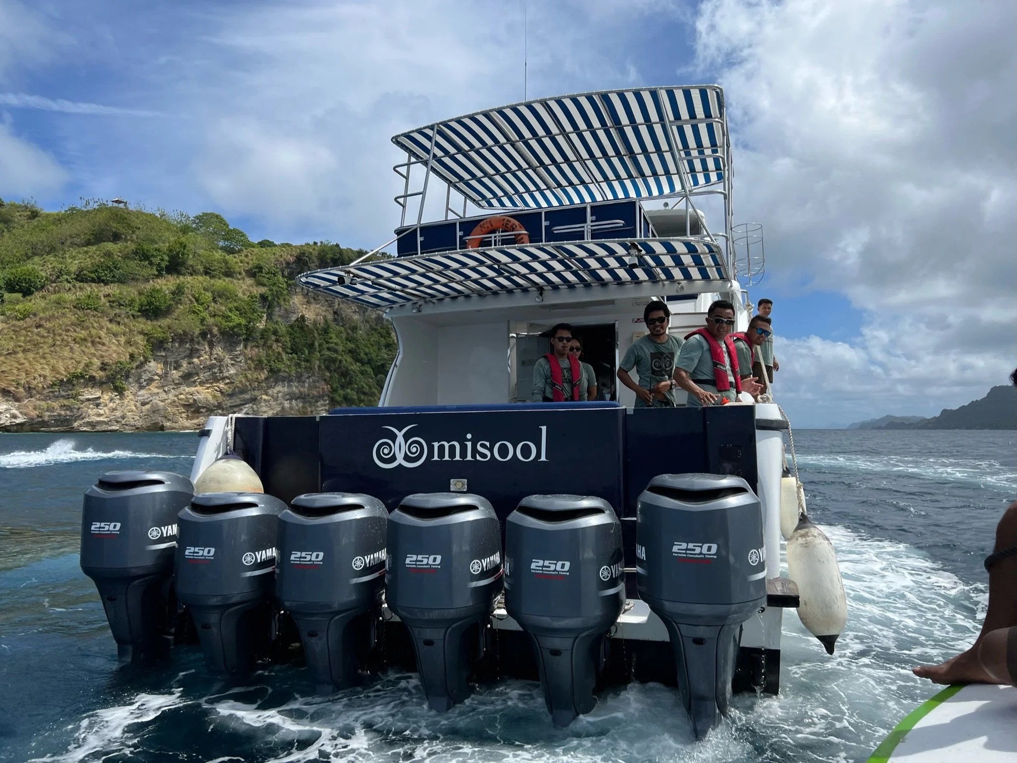 A group of five men on a boat with multiple Yamaha outboard engines, wearing life jackets, with a lush green hillside and a partly cloudy sky in the background.