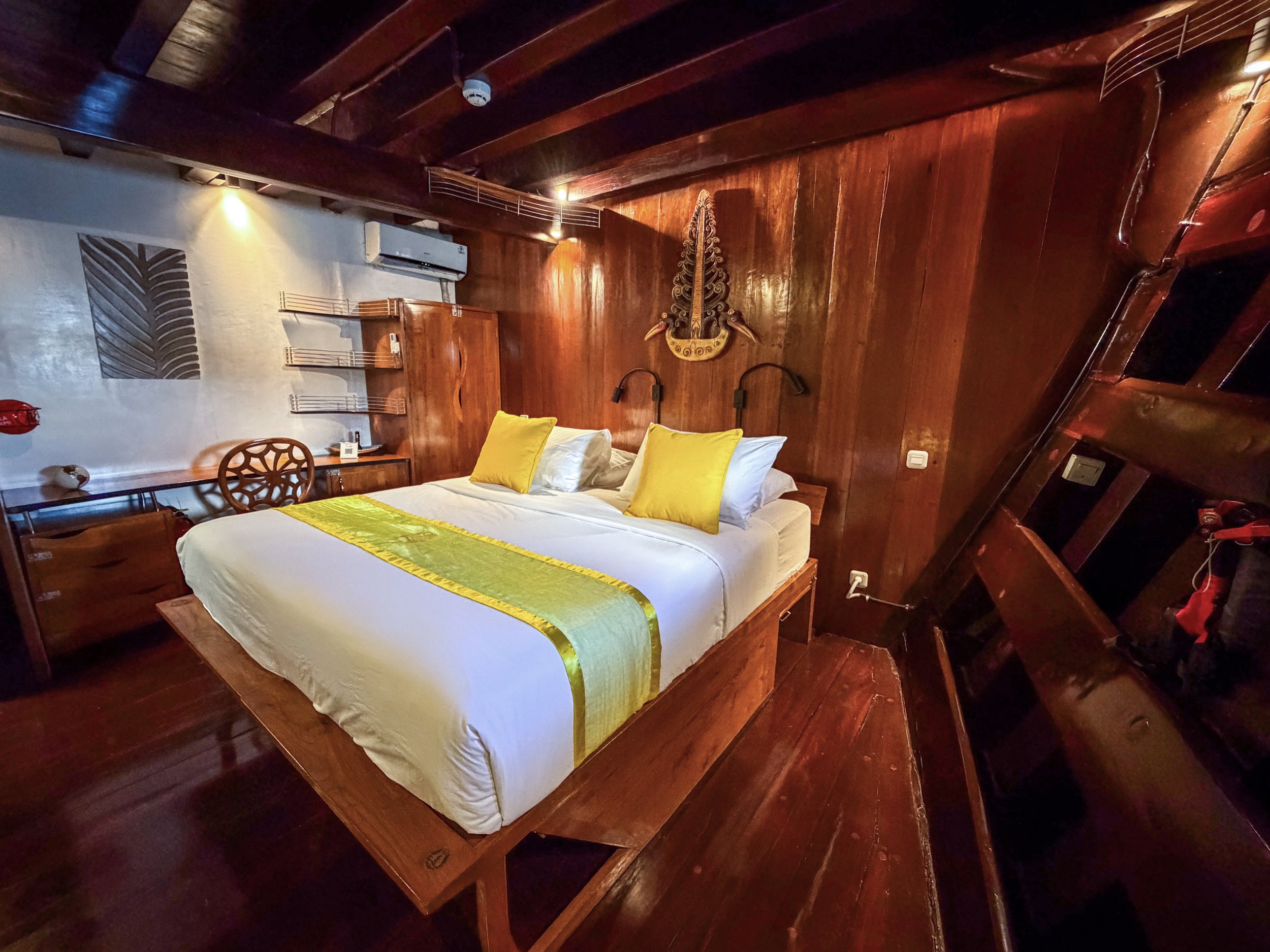 A bedroom with dark wood walls and ceiling, featuring a bed with white sheets, yellow and white pillows, and yellow and green bed runner. There is a decorative wall hanging, wall-mounted reading lights, a desk, a wicker chair, and a split air conditioning unit.