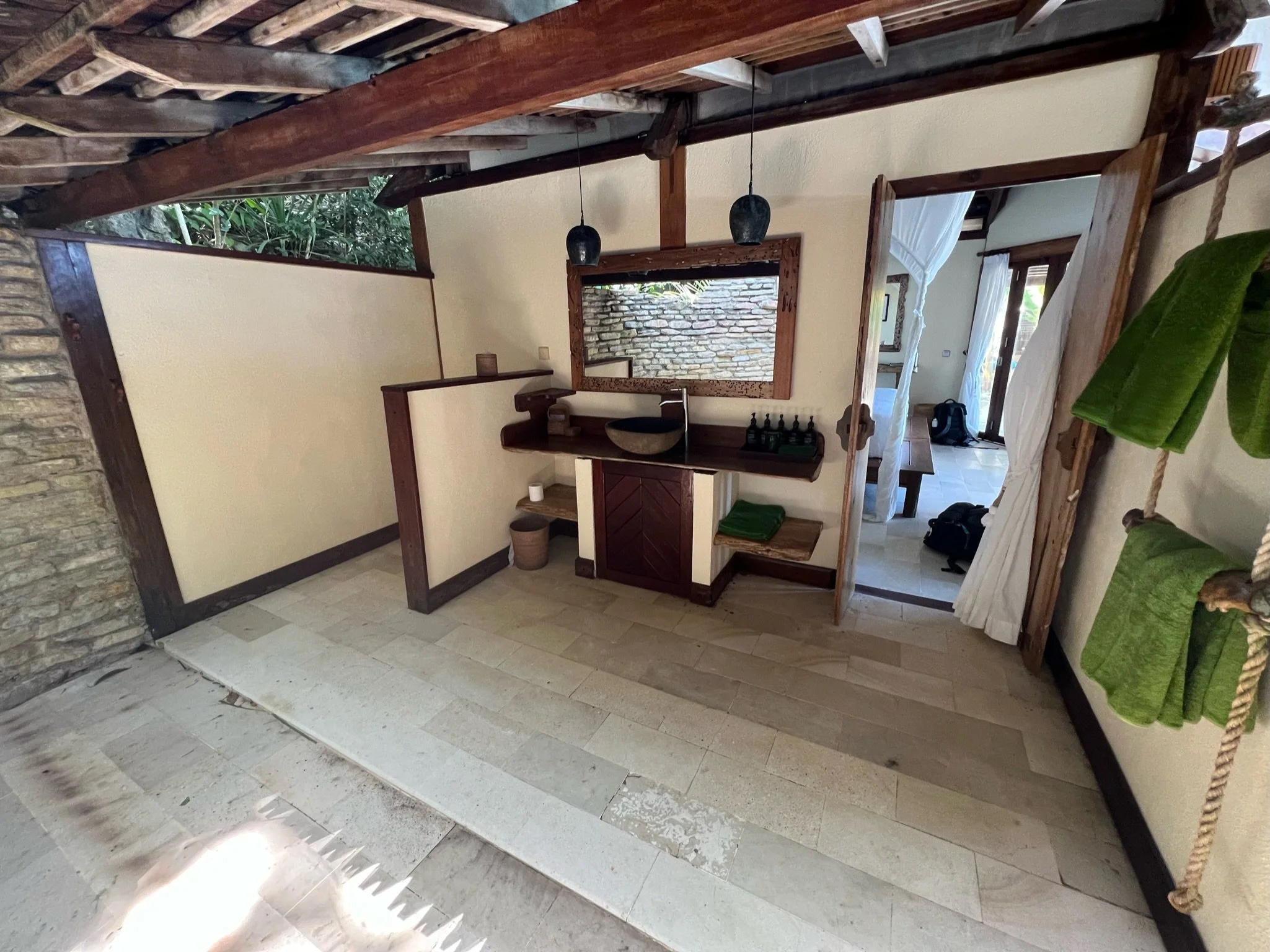 Rustic interior space with stone wall, wooden beams, hanging lights, and a small washbasin area with towels and toiletries, opening to an outdoor patio.