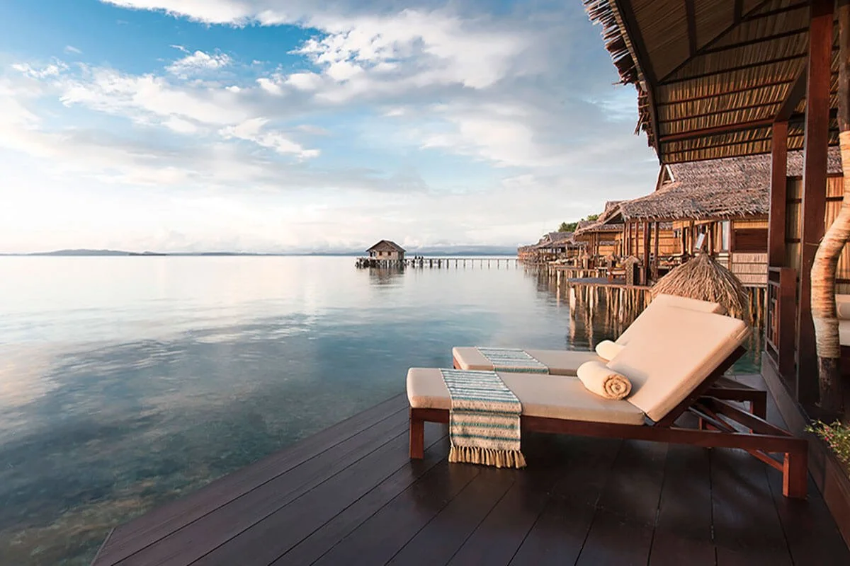 Overwater bungalow with a lounge chair and rolled towel on a wooden deck overlooking calm water and a distant hut on a pier, under a partly cloudy sky.