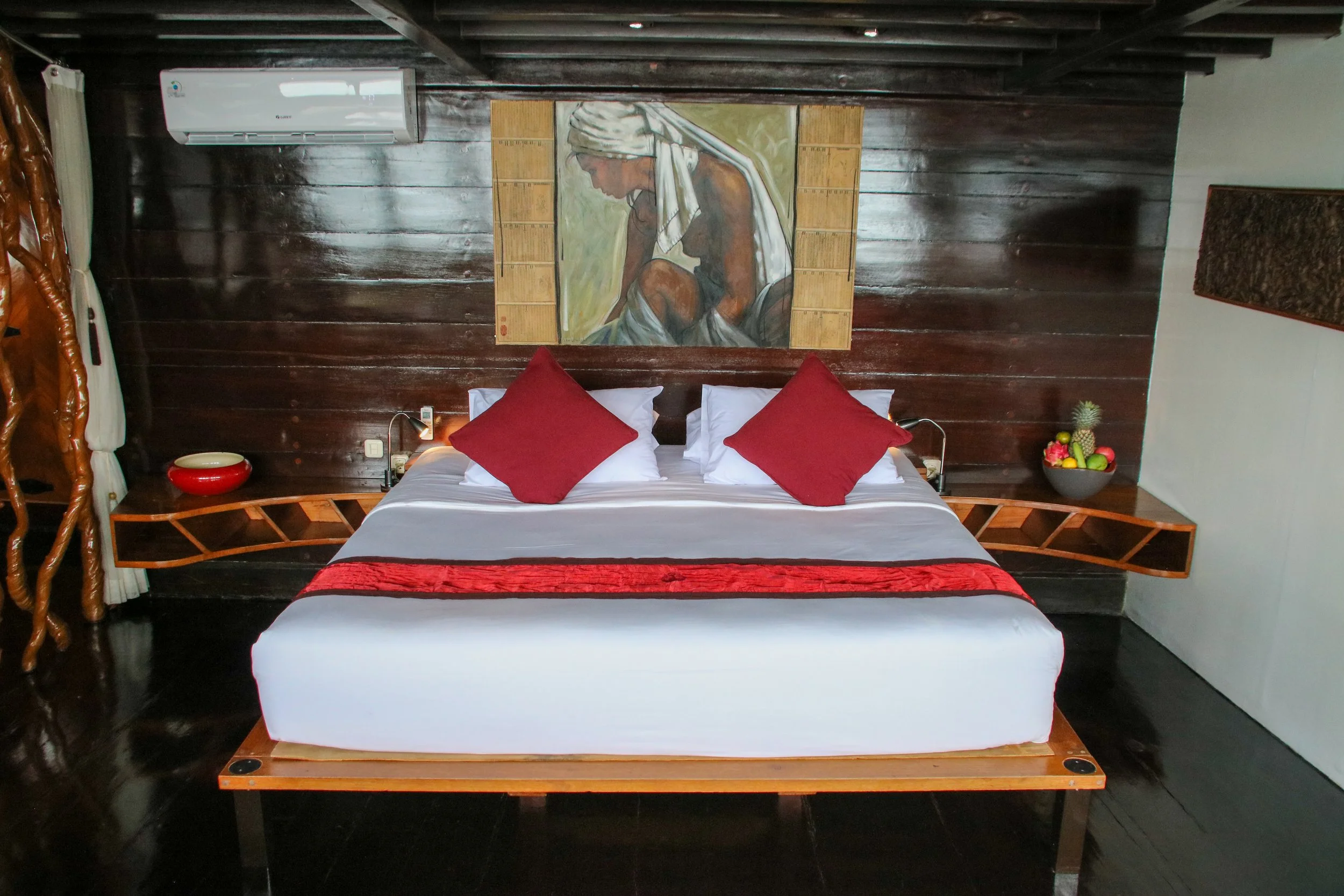 A bedroom with a dark wood accent wall behind a bed with white sheets, red pillows, and a red bed runner. Above the bed hangs a painting of a woman with a head covering. On each side of the bed are wooden side tables with lamps, a bowl of fruit on the right, and an air conditioning unit on the wall.