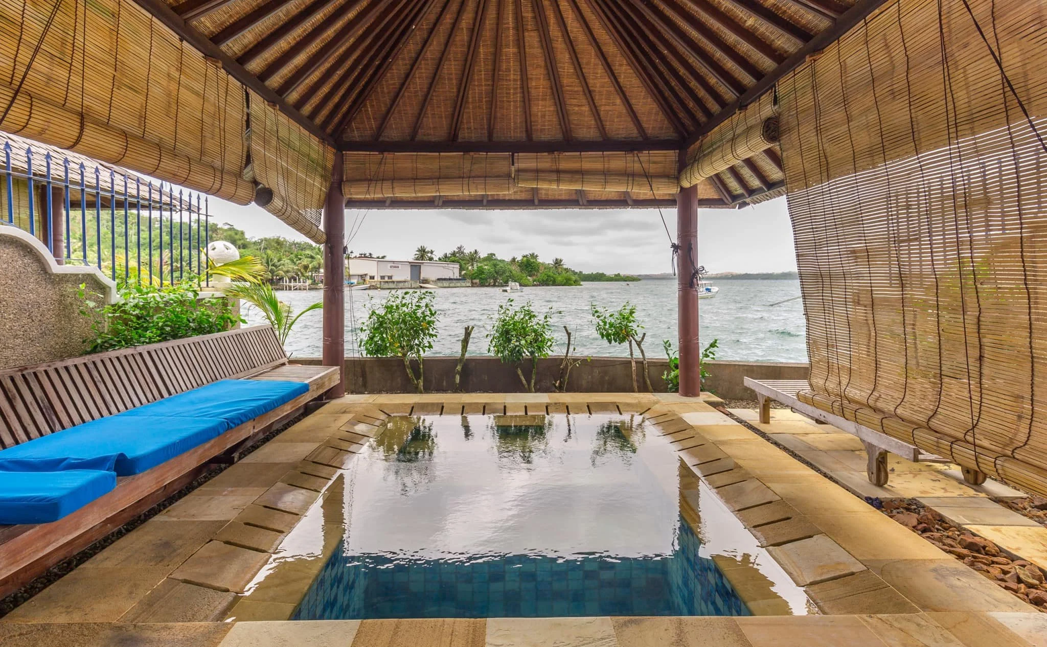 Open-air poolside cabana with bamboo roof, lakeside view, wooden benches with blue cushions, and nearby greenery.