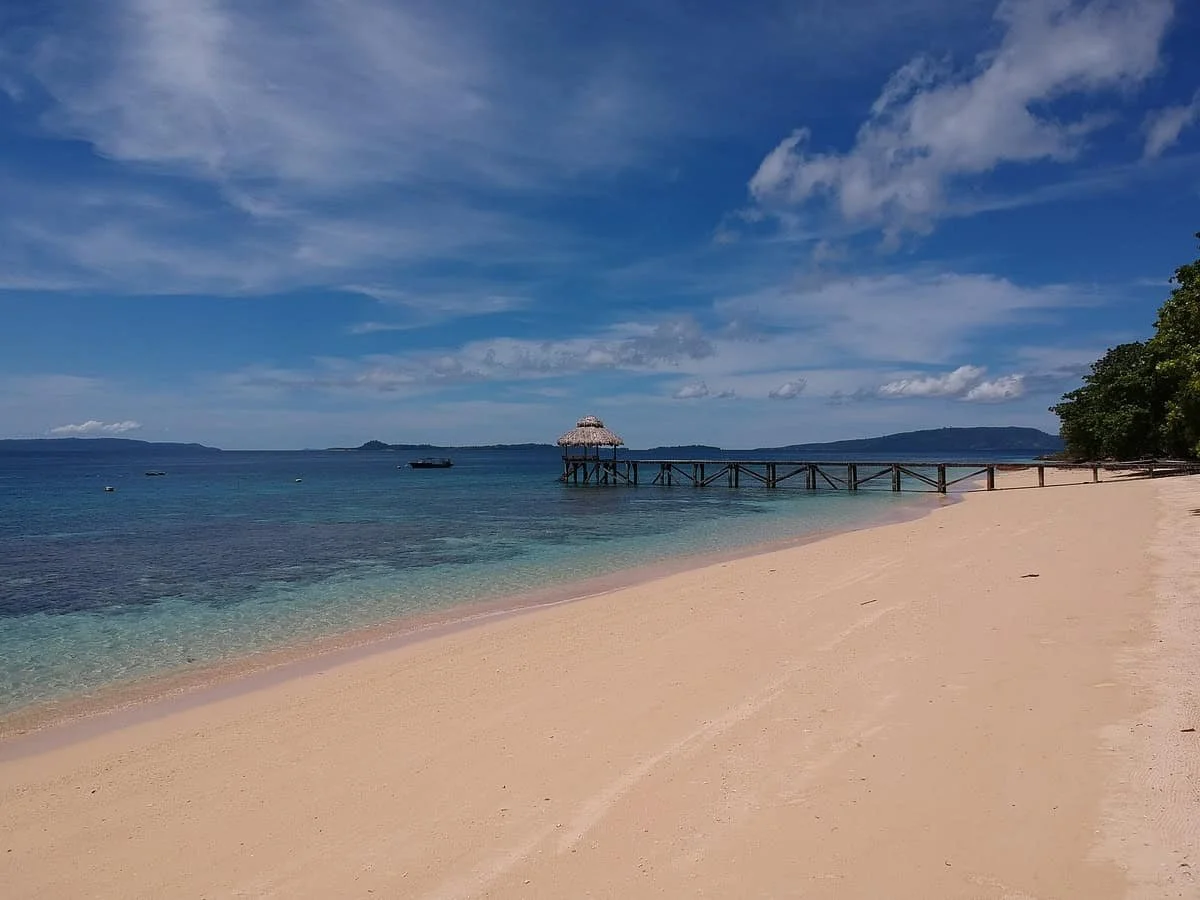 Sunny beach with a wooden pier and a thatched-roof hut extending over calm blue water, with distant islands on the horizon, partly cloudy sky, and trees on the right side of the beach.
