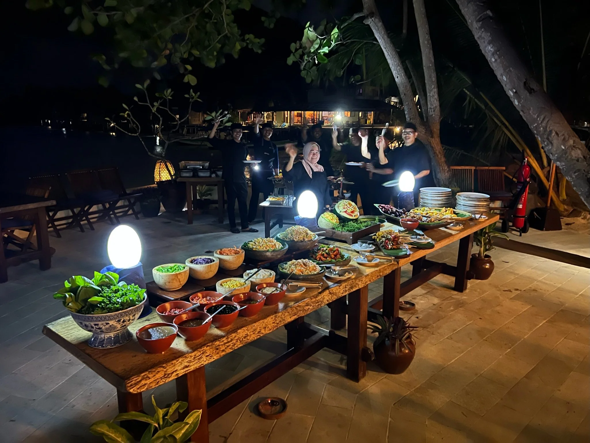 Nighttime outdoor buffet table with various dishes, with a group of smiling staff members waving in the background, surrounded by trees and seating area.