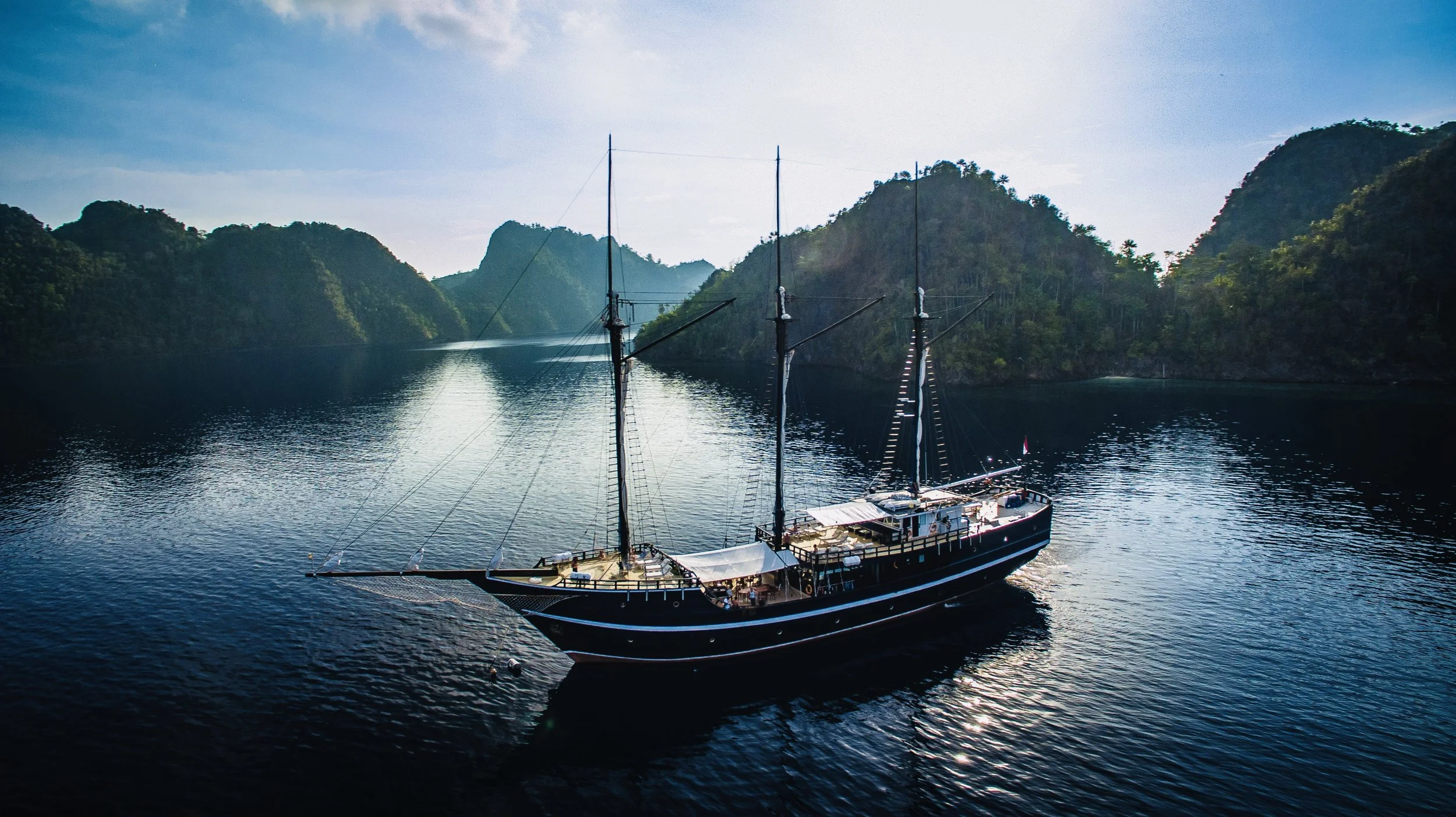 A black and white sailing ship with multiple masts anchored in a calm body of water, surrounded by green forested hills and mountains under a clear blue sky.