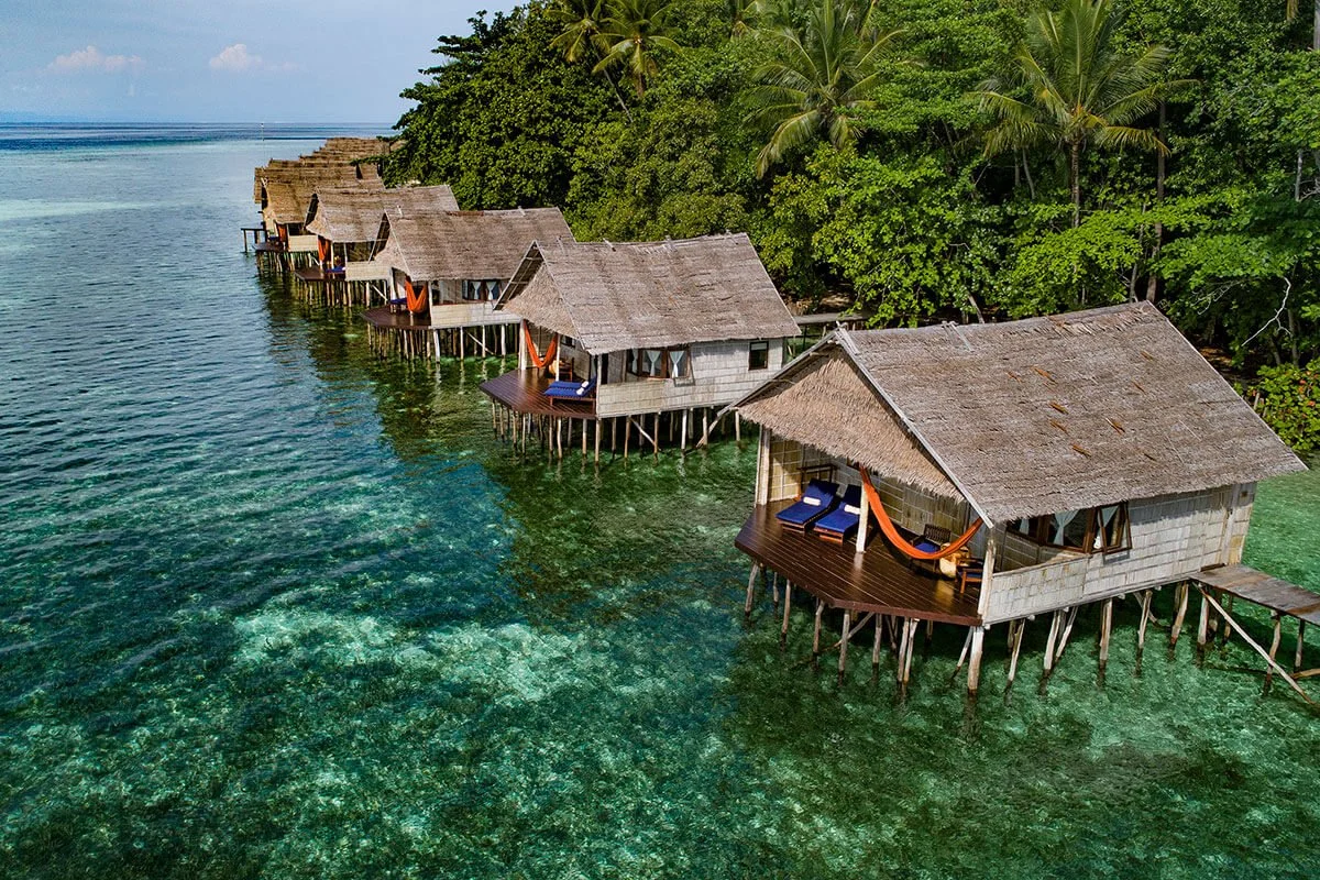 Overwater bungalows with thatched roofs and wooden decks, hammock, and lounge chairs, situated over clear, turquoise ocean water, with lush green trees in the background.