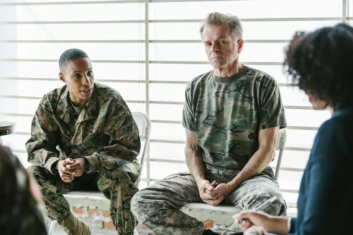 A military woman and a man wearing camouflage clothing sitting during a discussion with two women, one with curly hair, in a room with bright windows.