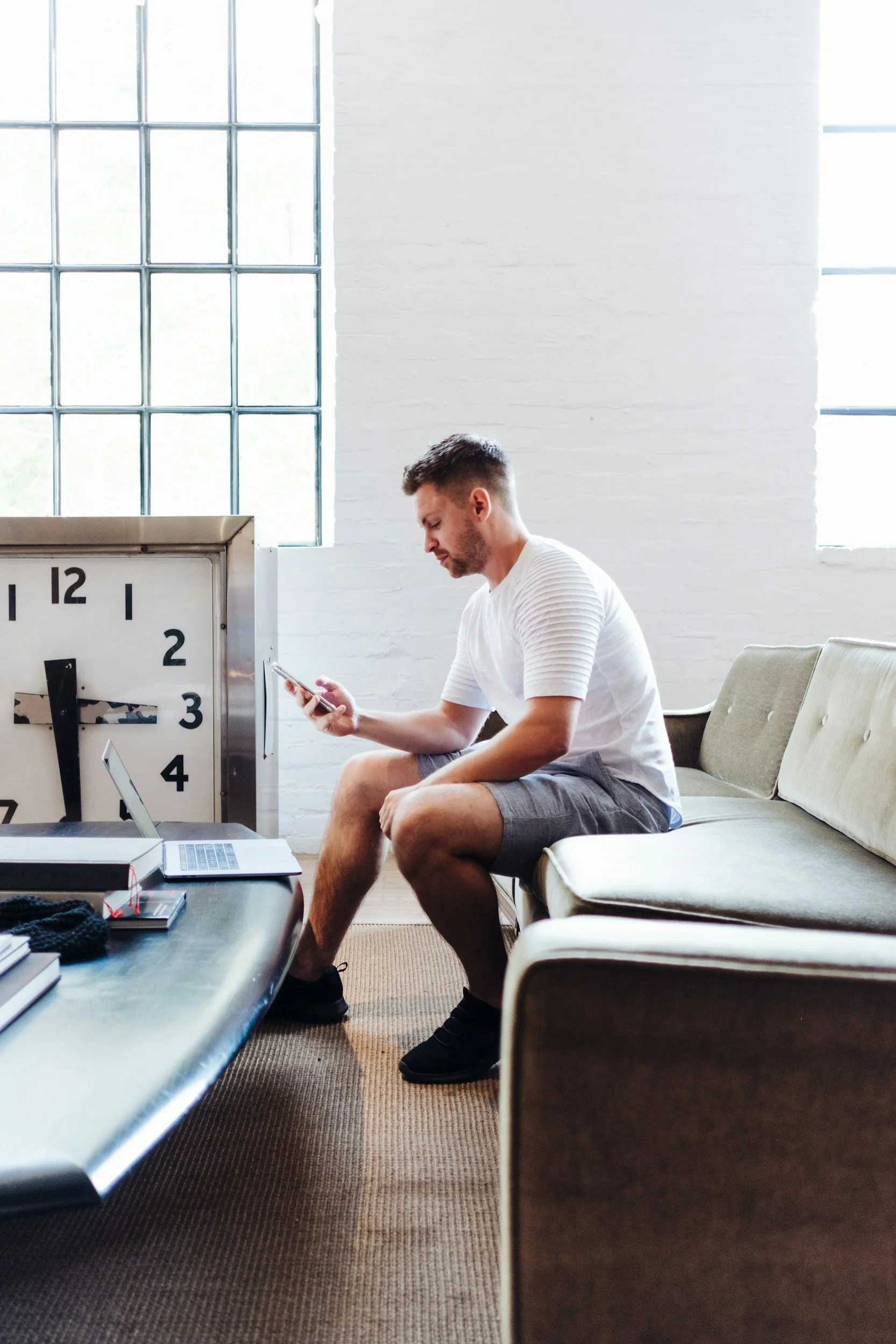 Man in white T-shirt and shorts sitting on a beige sofa, looking at his phone, near a large clock, in a bright therapy room with large windows.