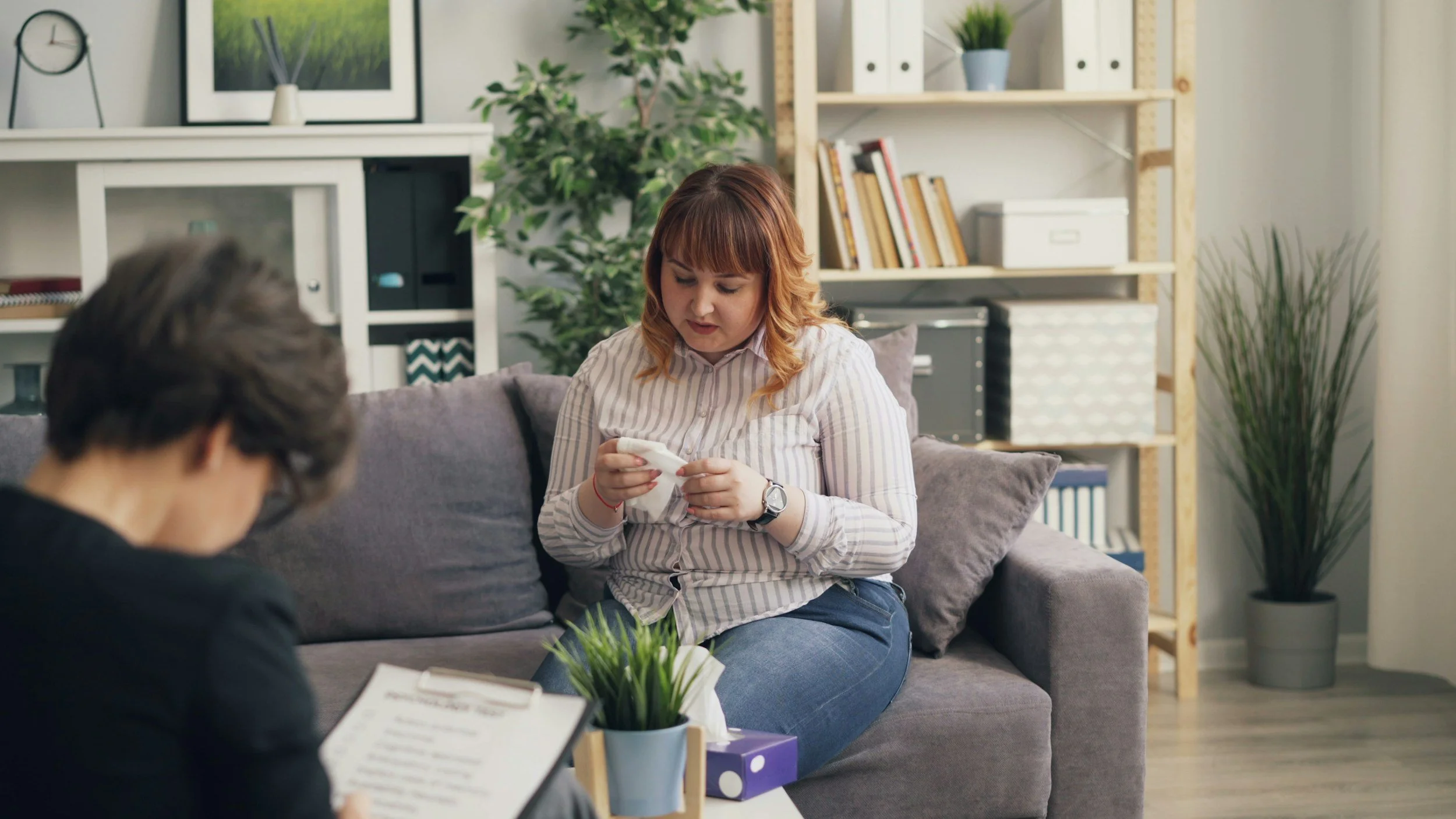 A woman with reddish-brown hair and a striped shirt sitting on a sofa, looking down at her hands, in a cozy therapy room with bookshelves, plants, and framed artwork.