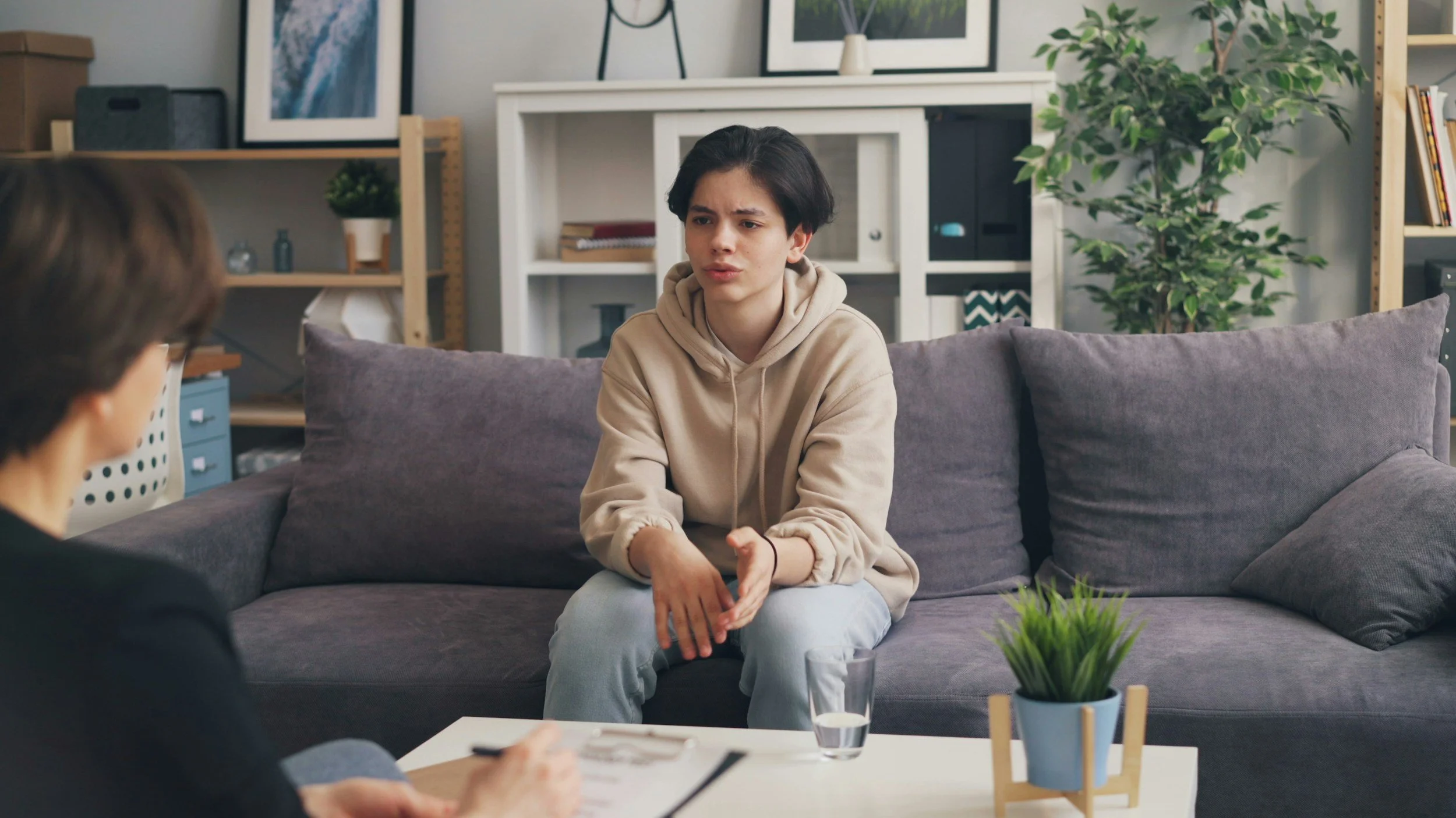 A young woman with dark hair and a beige hoodie sitting on a grey couch, appearing to speak during a conversation in a therapy room with artwork, plants, and shelves in the background.