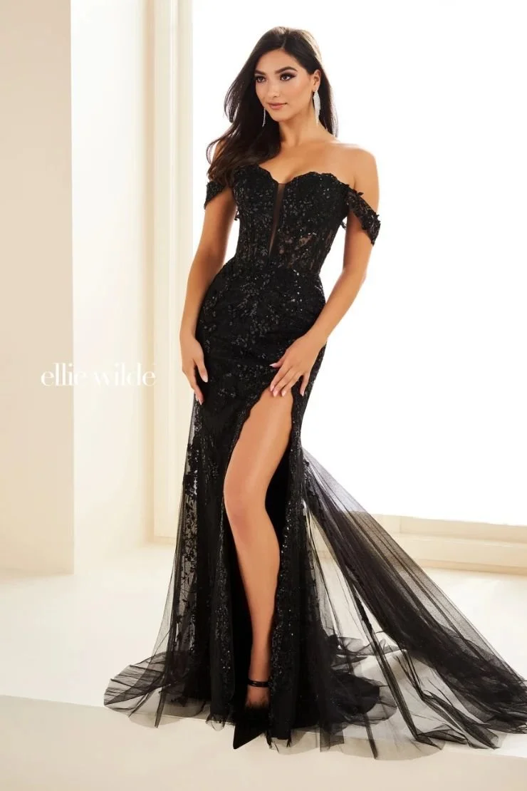 Woman wearing a black, off-shoulder, sequined evening gown with a high slit, standing in a well-lit room.