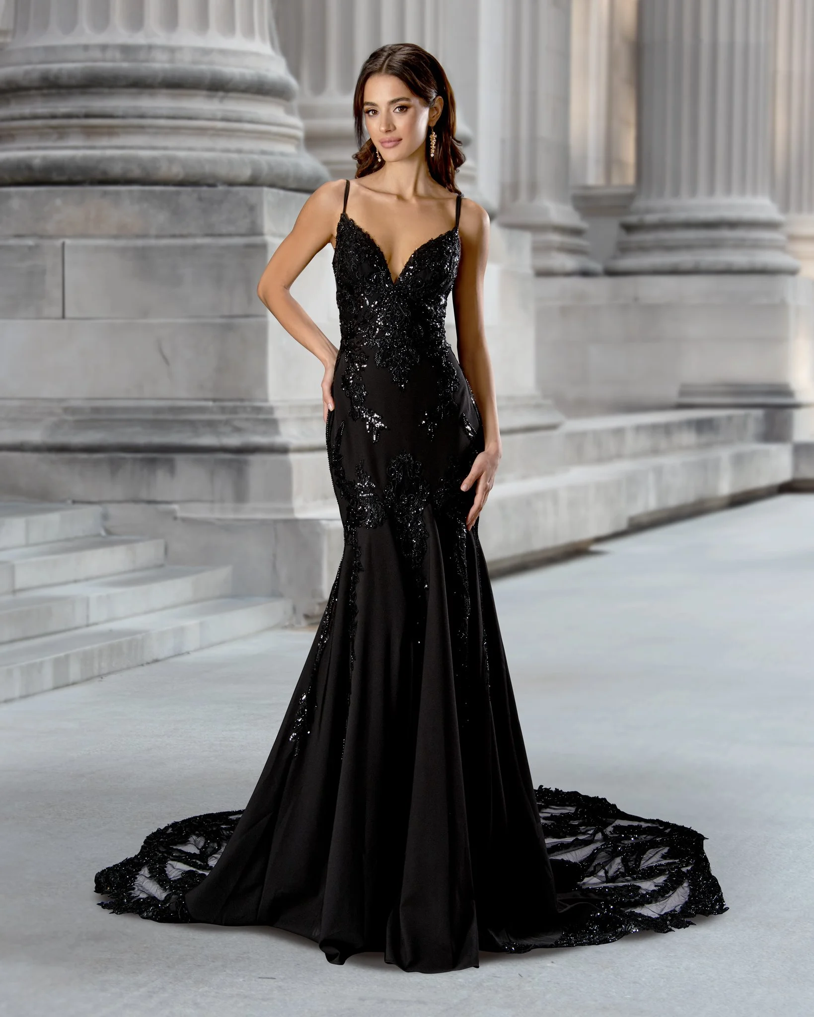 A woman in a fitted black gown with sequins, lace details, and a train, standing in front of stone classical columns. She has dark, shoulder-length hair and is wearing long earrings.