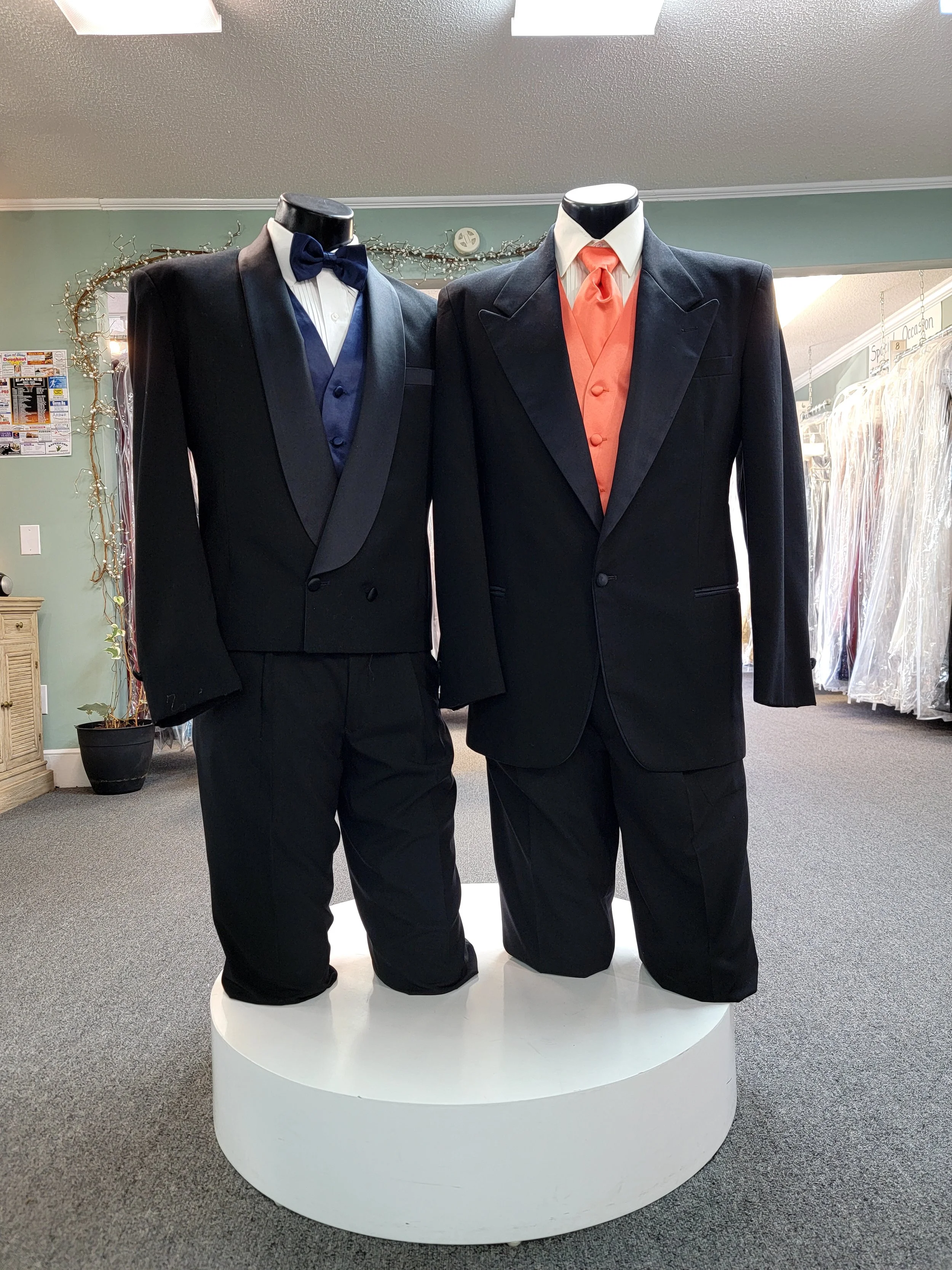 Two tuxedo mannequins displaying formal suits, one with a navy blue tuxedo with a bow tie, and the other with a black tuxedo with an orange vest and tie, in a retail store setting.