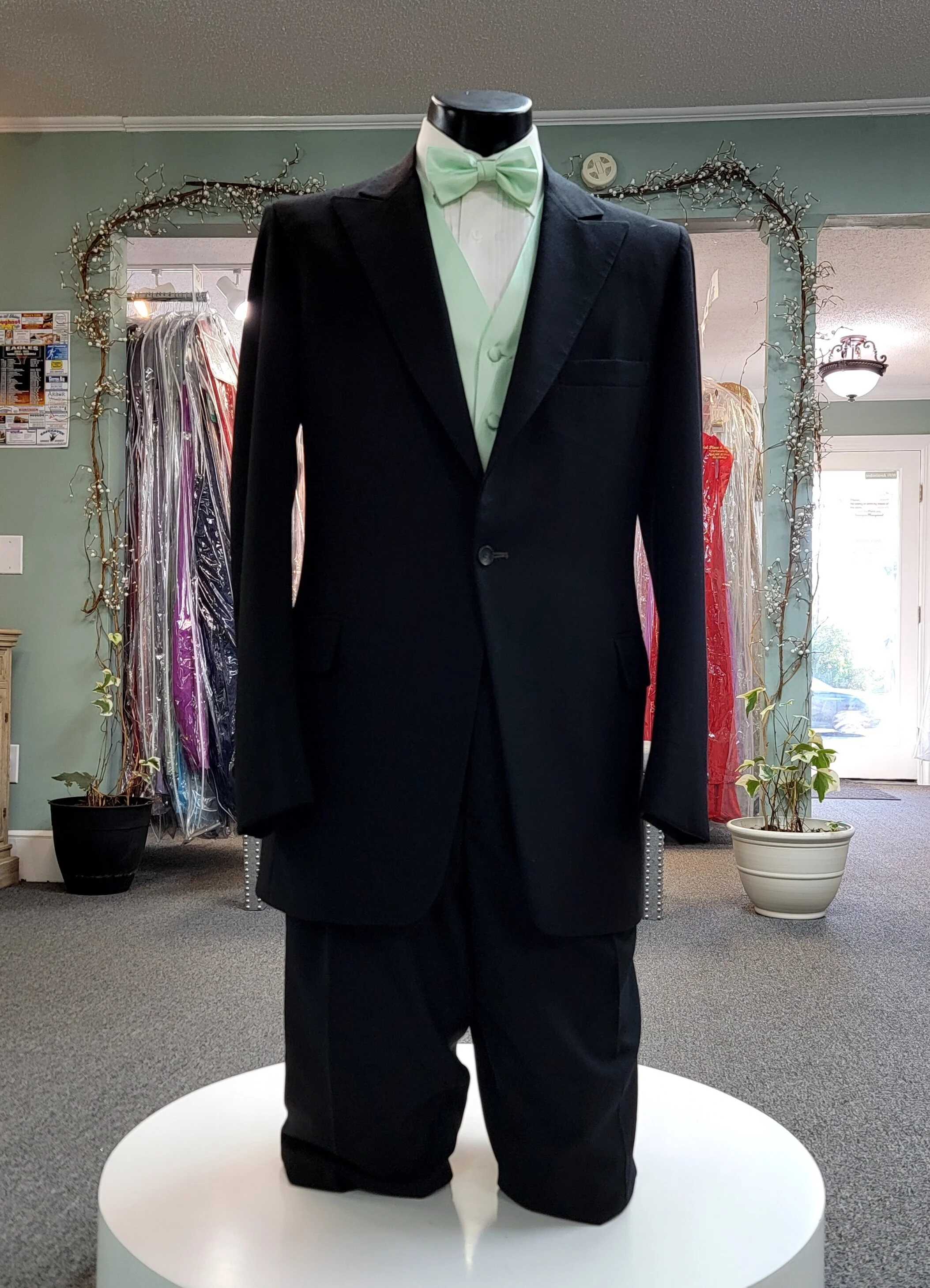 Male mannequin wearing a black tuxedo with a mint green bow tie and vest, displayed on a white round platform inside a store with clothing racks, potted plants, and decorative lights.