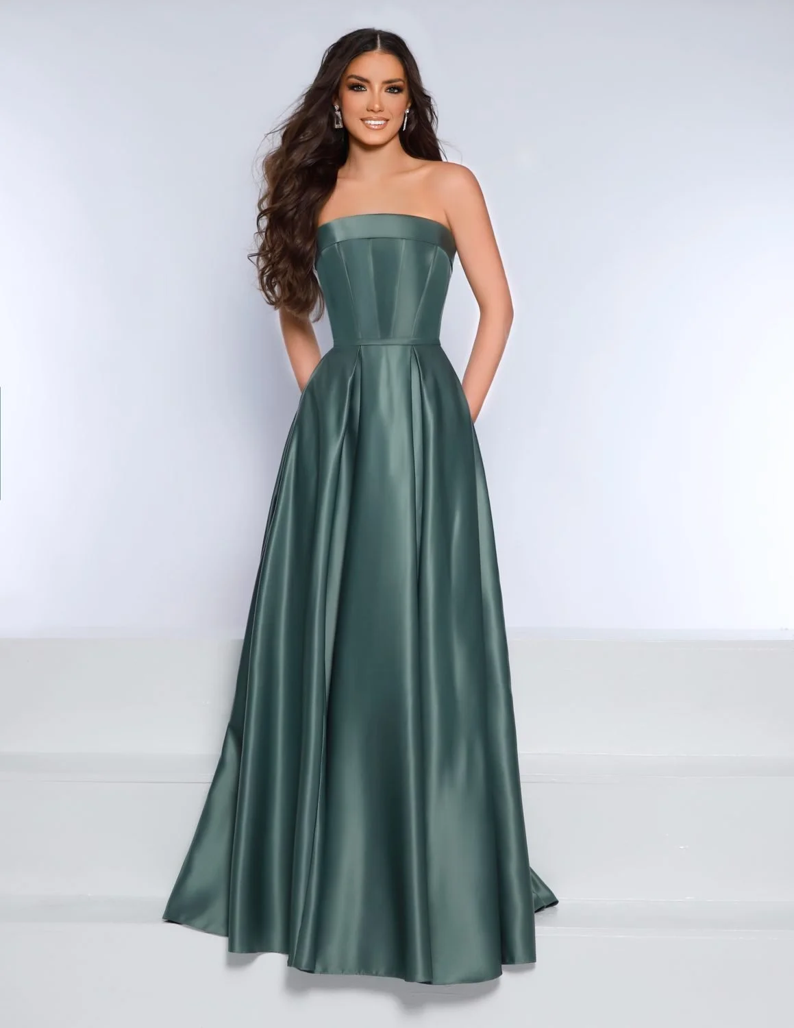 A woman with long, dark wavy hair wearing a strapless green satin evening gown, standing against a plain light gray background.