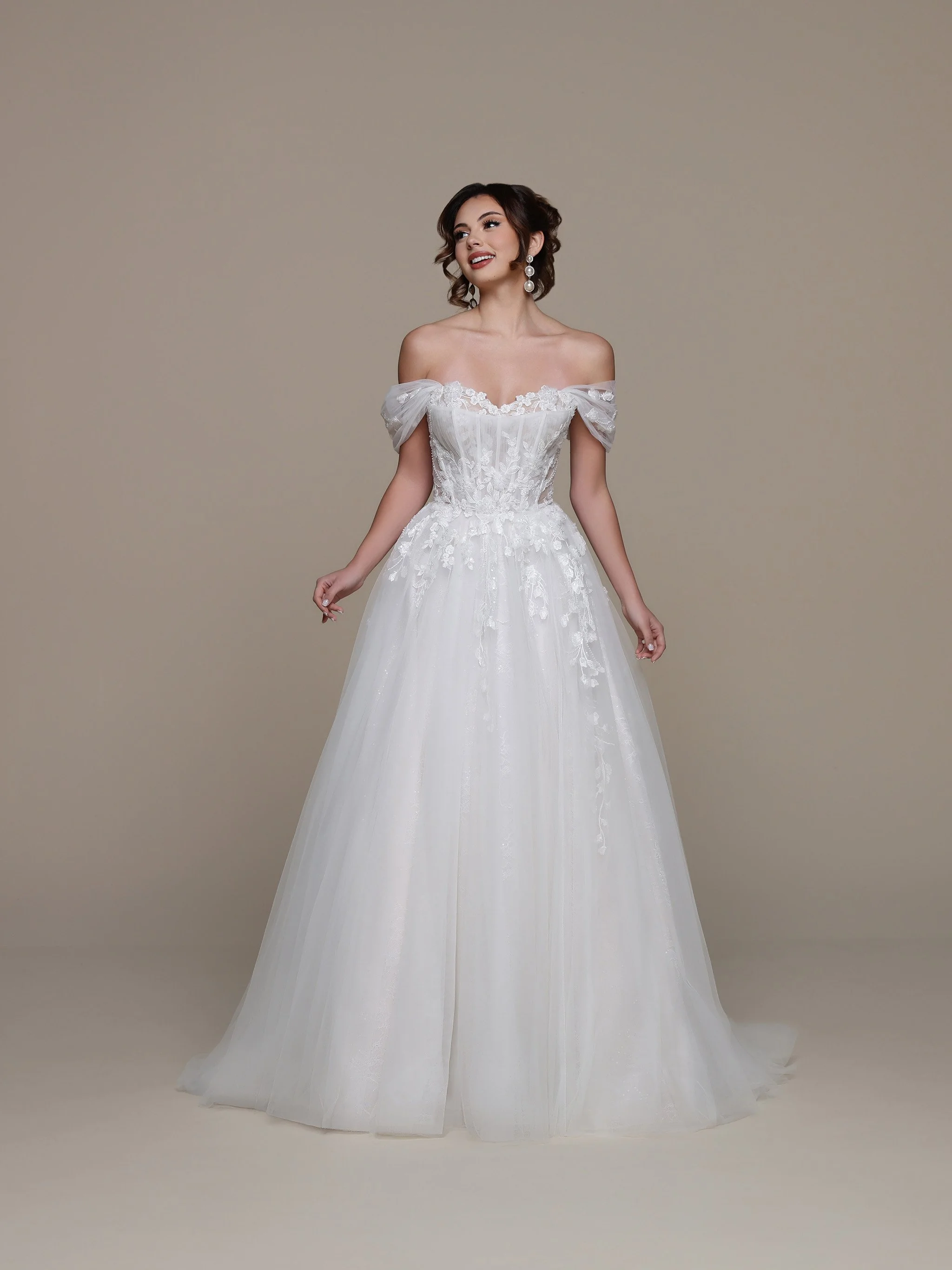 A woman wearing a white off-shoulder wedding dress with lace details, standing against a plain beige background.